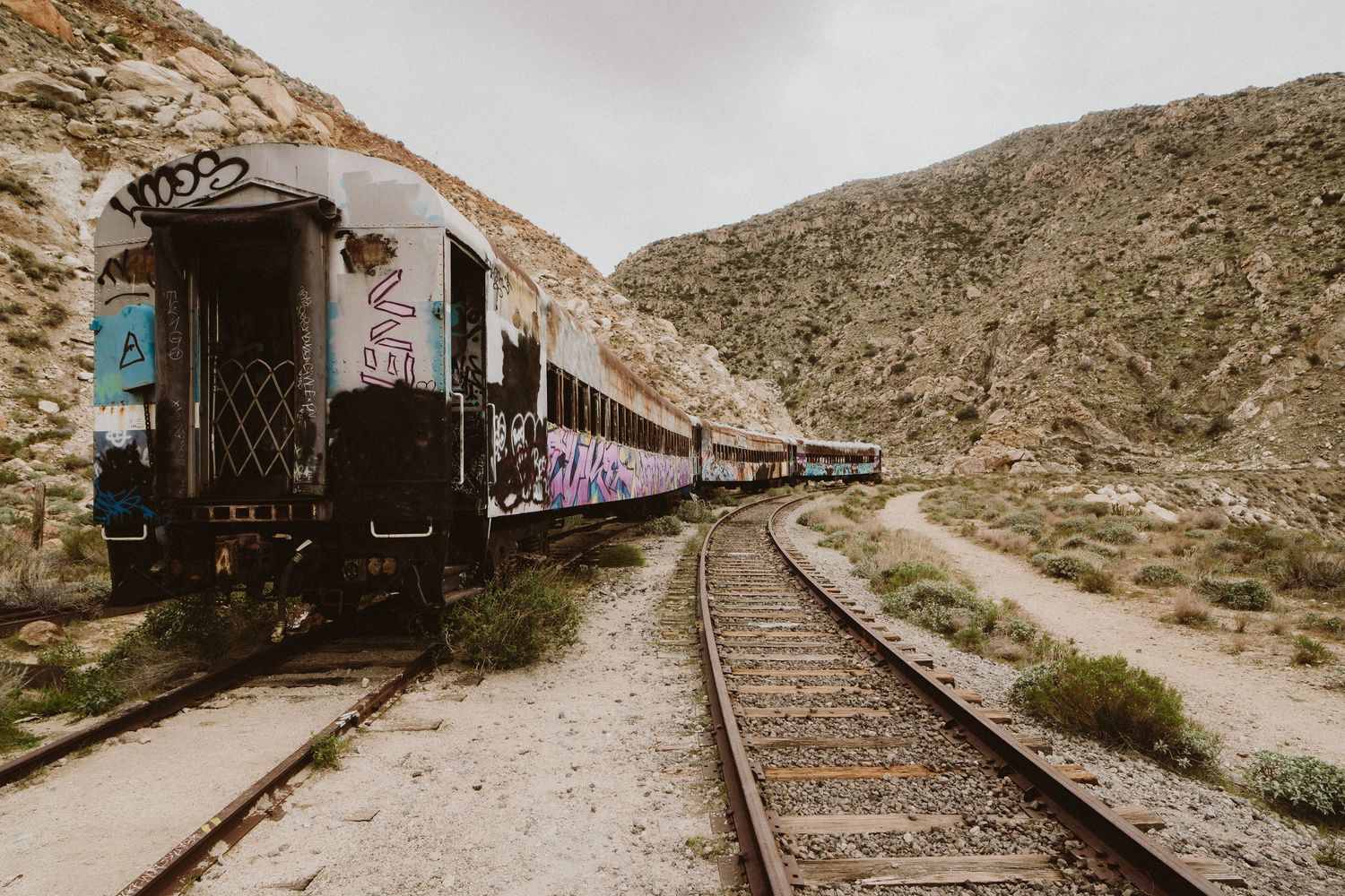 Vintage passenger train cars parked on railroad tracks winding through rugged desert mountains in muted tones.