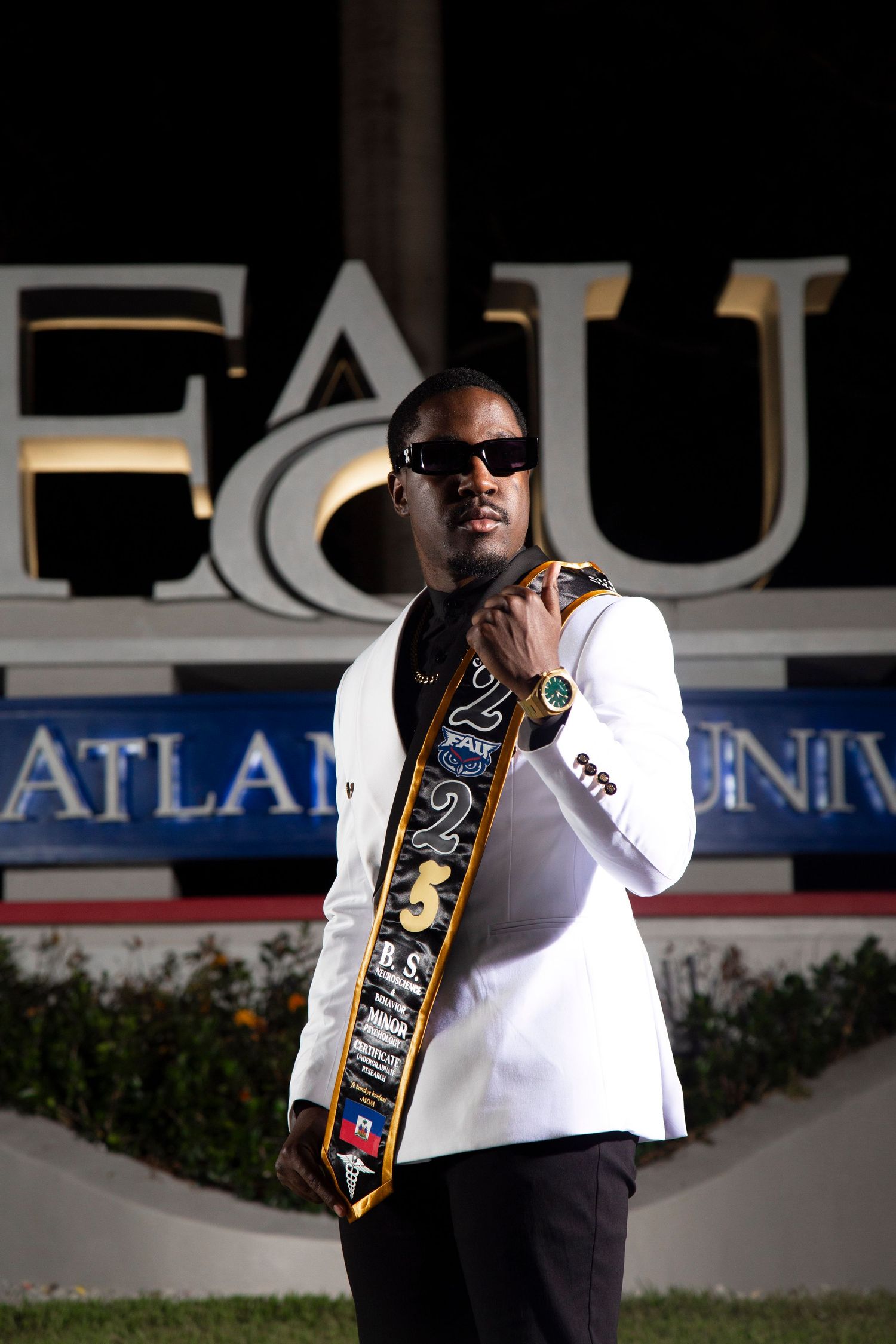 Graduate wearing sunglasses and graduation stole poses in front of university signage.
