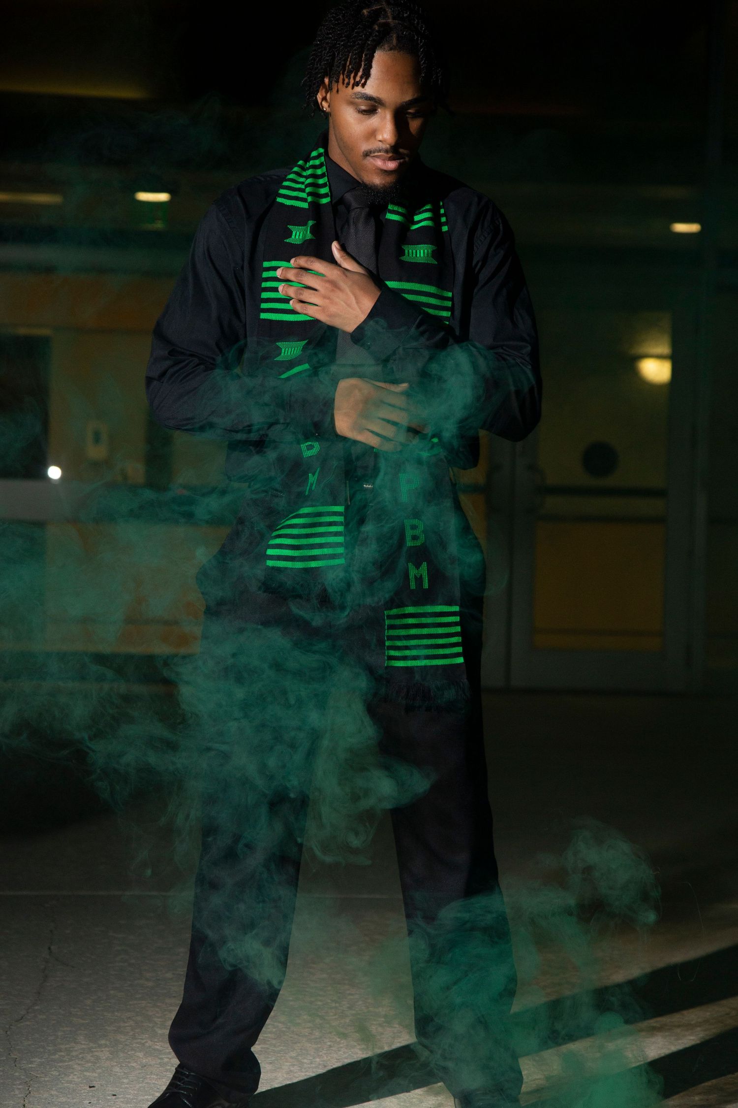 Person in dark clothing with green stripes poses in atmospheric green smoke effects in a dimly lit indoor space.
