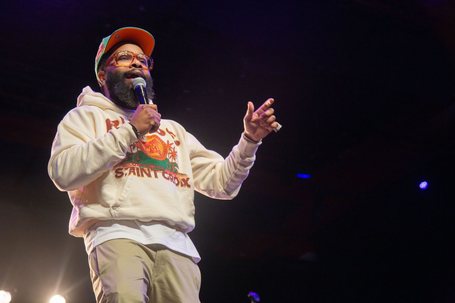 Performer in orange cap and cream hoodie gestures while speaking on stage with blue lights in background.
