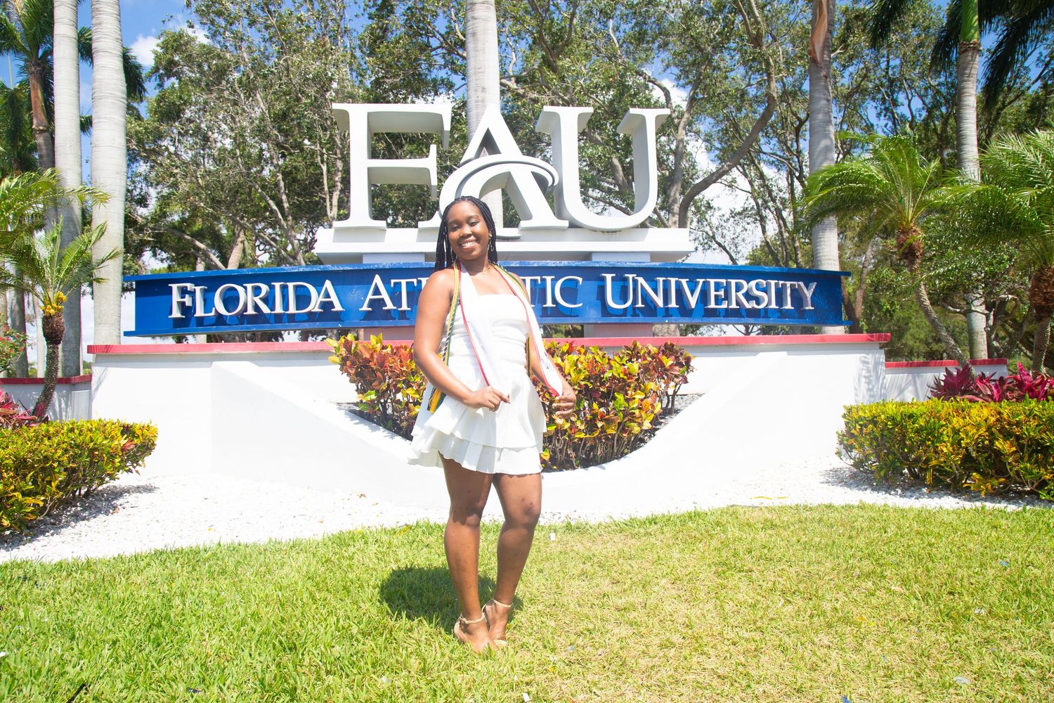 Graduate poses in front of Florida Atlantic University entrance sign surrounded by palm trees and colorful flowers.
