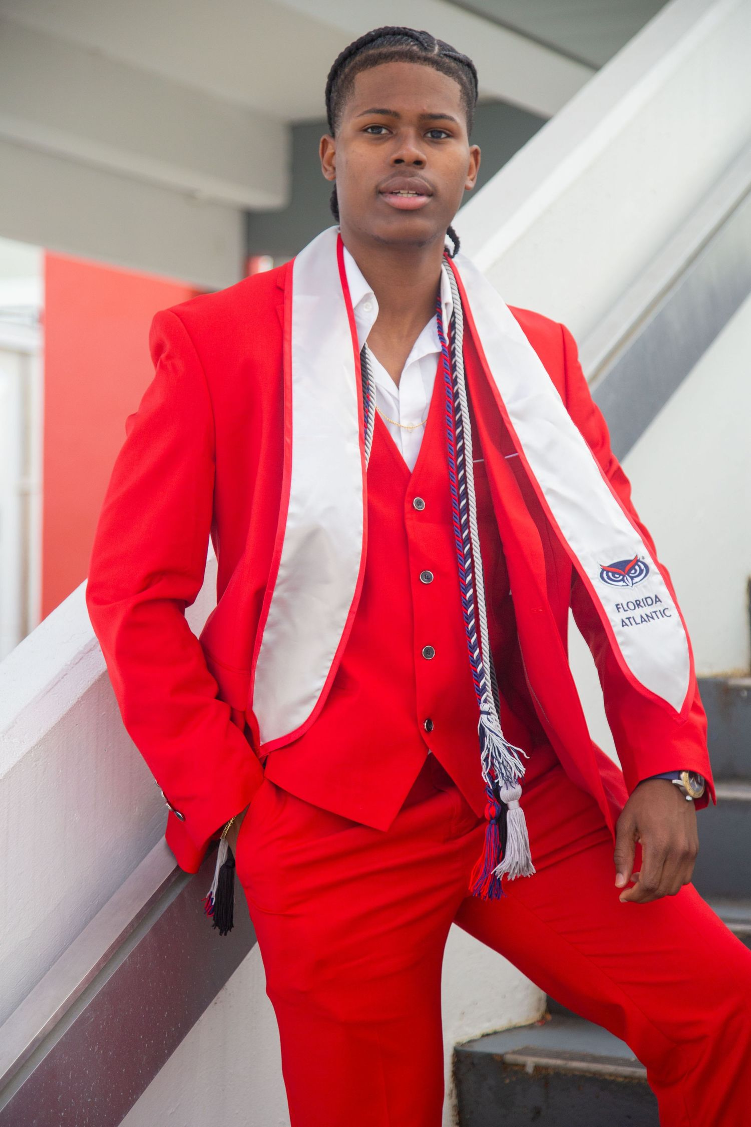 Graduate wearing striking red three-piece suit poses against white architectural background.