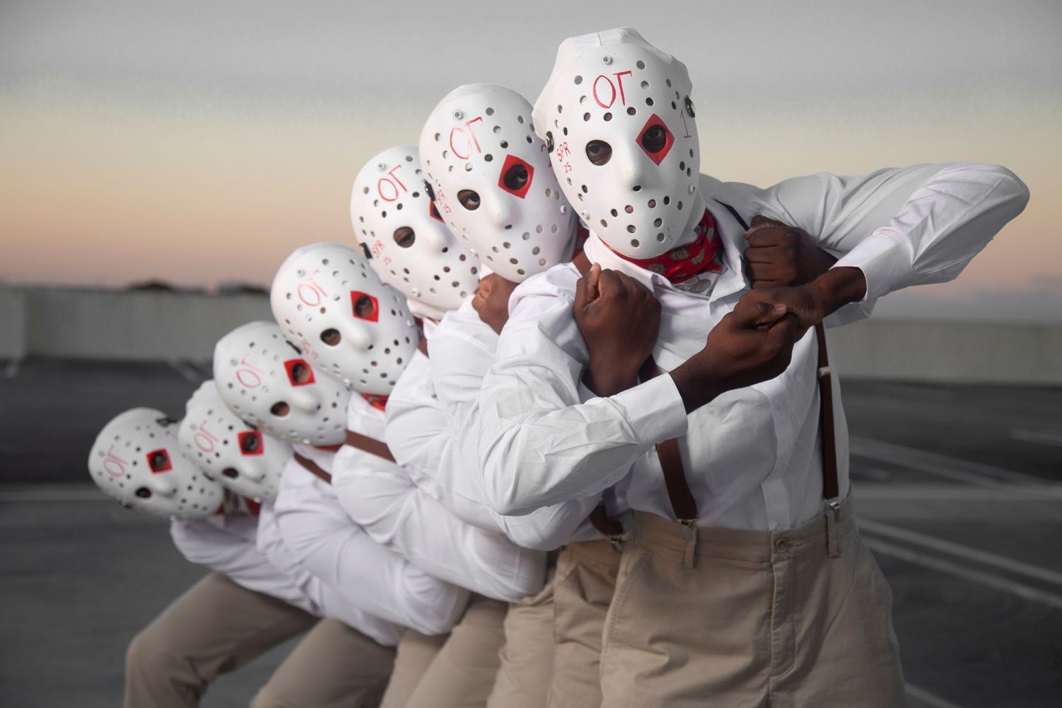 A row of performers wearing identical white hockey masks and outfits posing against a sunset background.