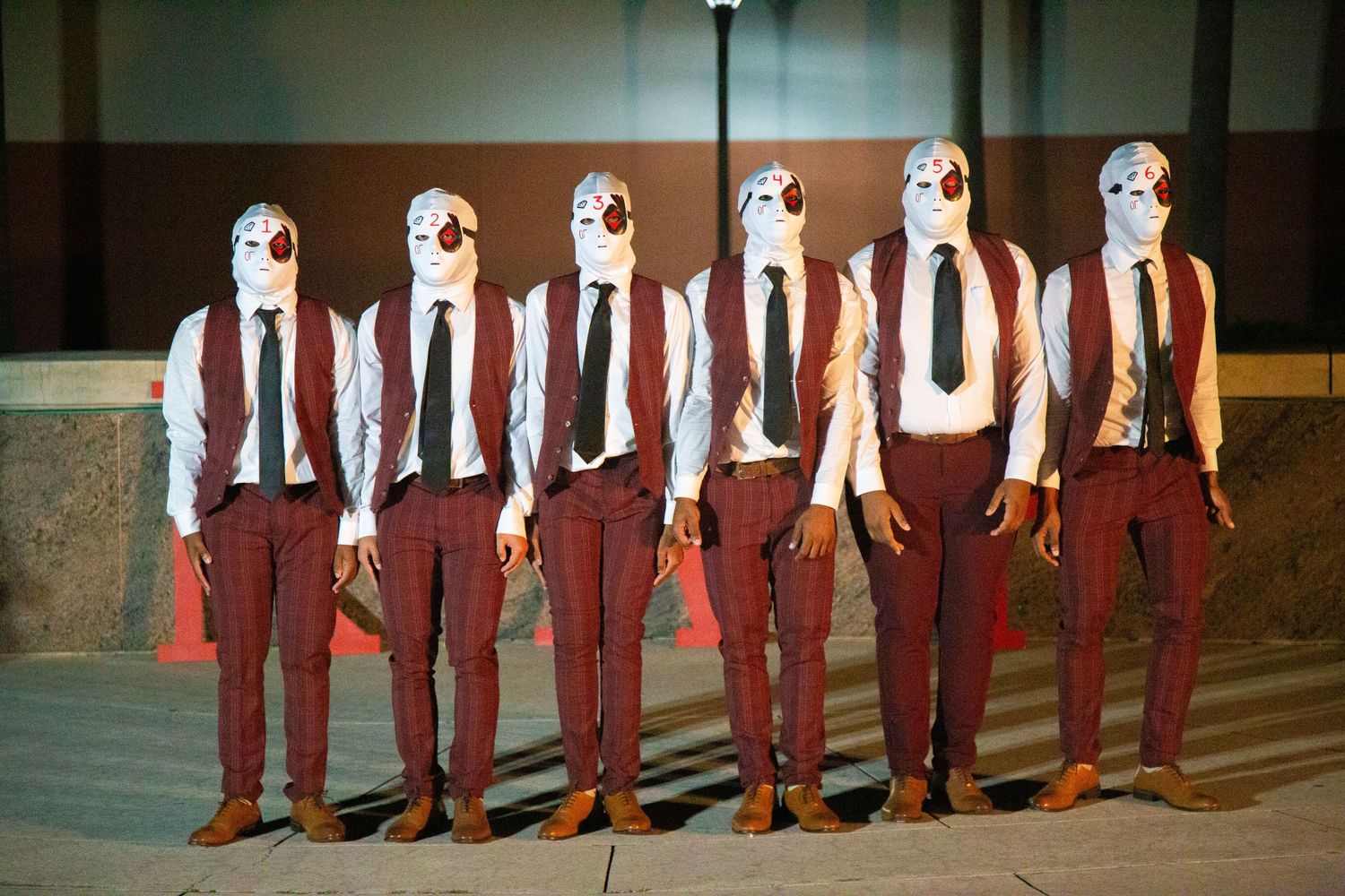 Six performers in white masks, red suits and ties stand in line on stage.