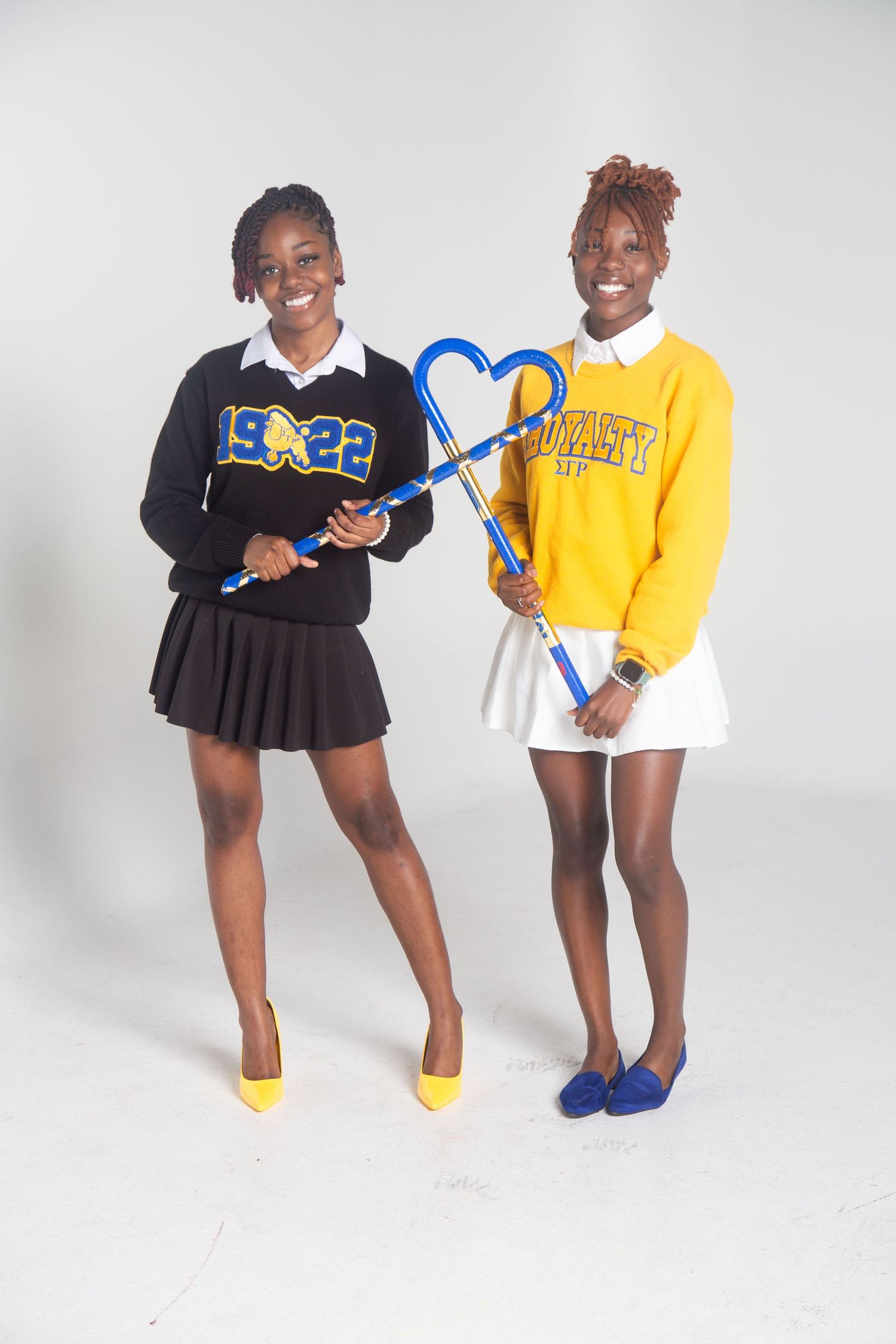 Two students in sorority sweaters and pleated skirts pose with blue decorative props against white backdrop.