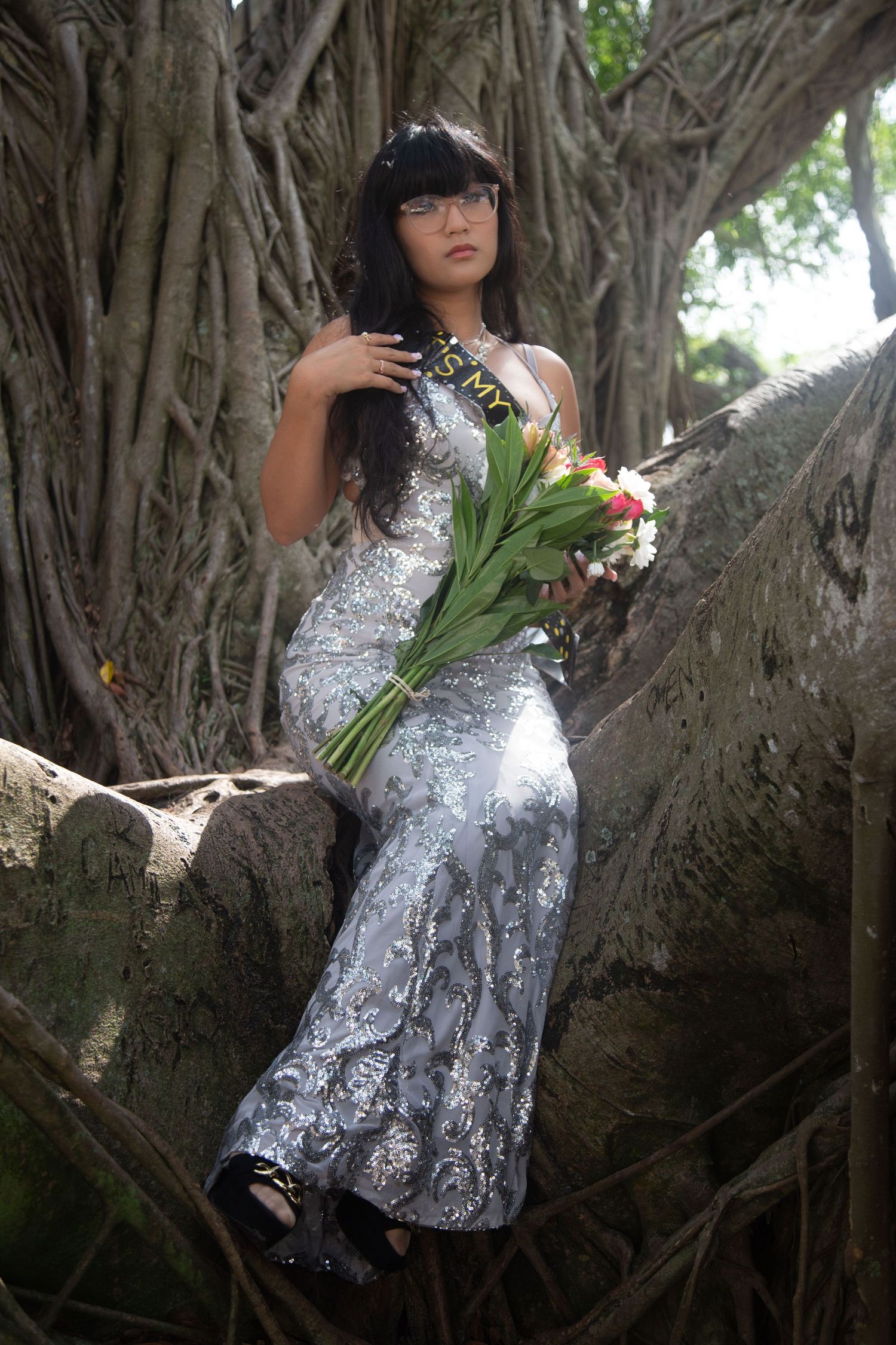 Person in formal silver dress sits gracefully in tree holding fresh flower bouquet.