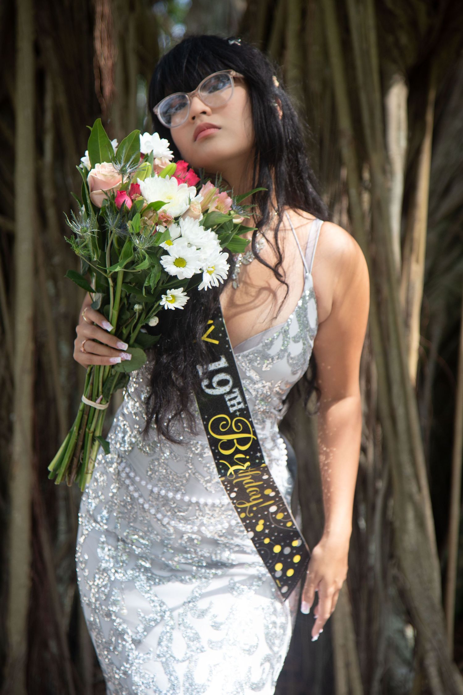 Woman in sparkly silver dress holds bouquet wearing birthday sash among bamboo trees.