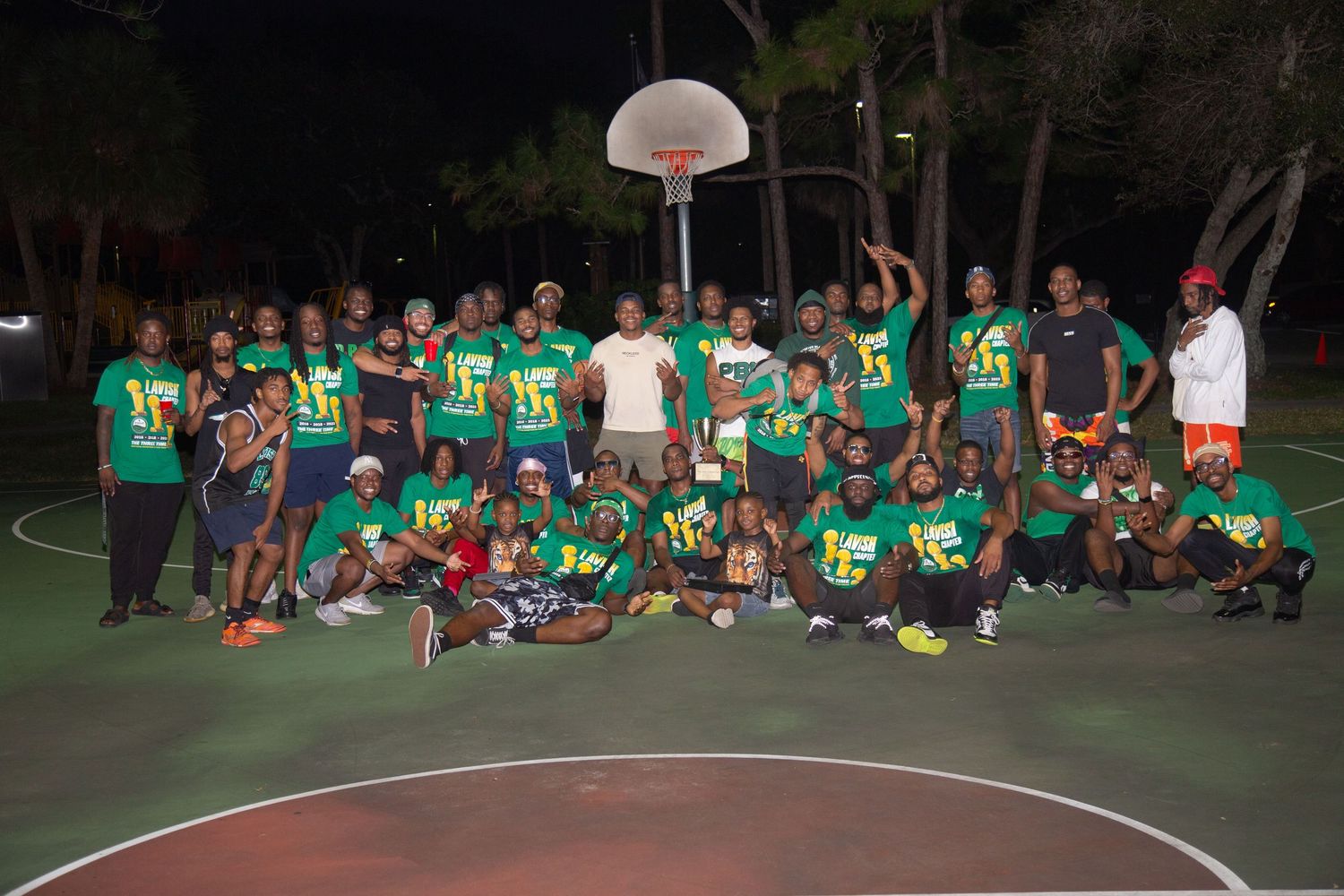 Large group in green jerseys poses on outdoor basketball court at night with hoop in background.