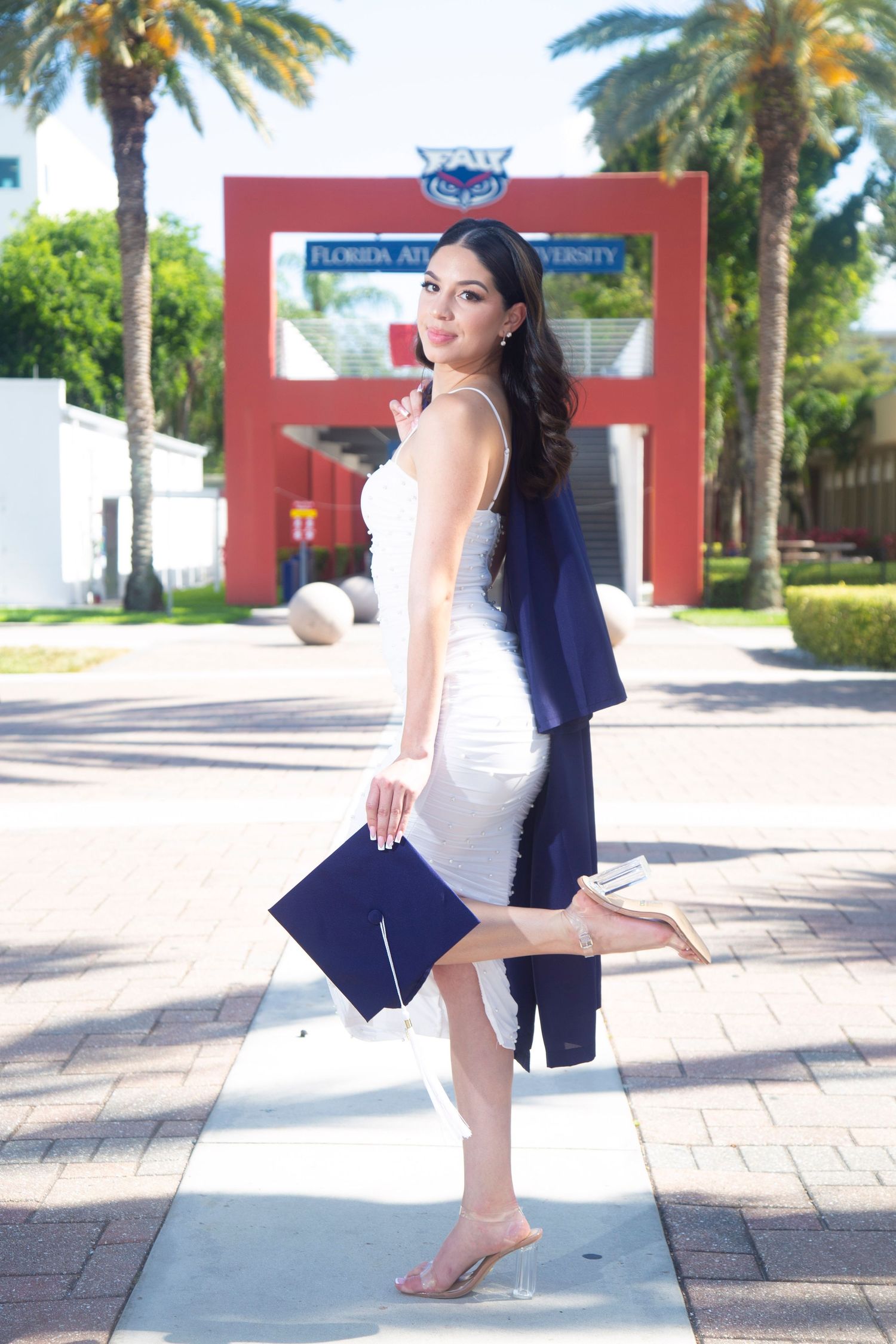 A graduate celebrates in a white dress and blue graduation cap while posing with palm trees in the background.