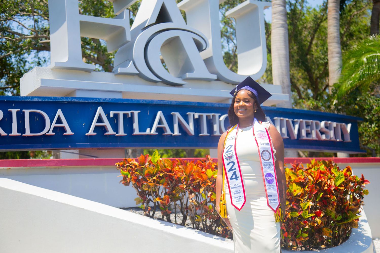 Graduate wearing honors sash poses in front of Florida Atlantic University entrance sign.