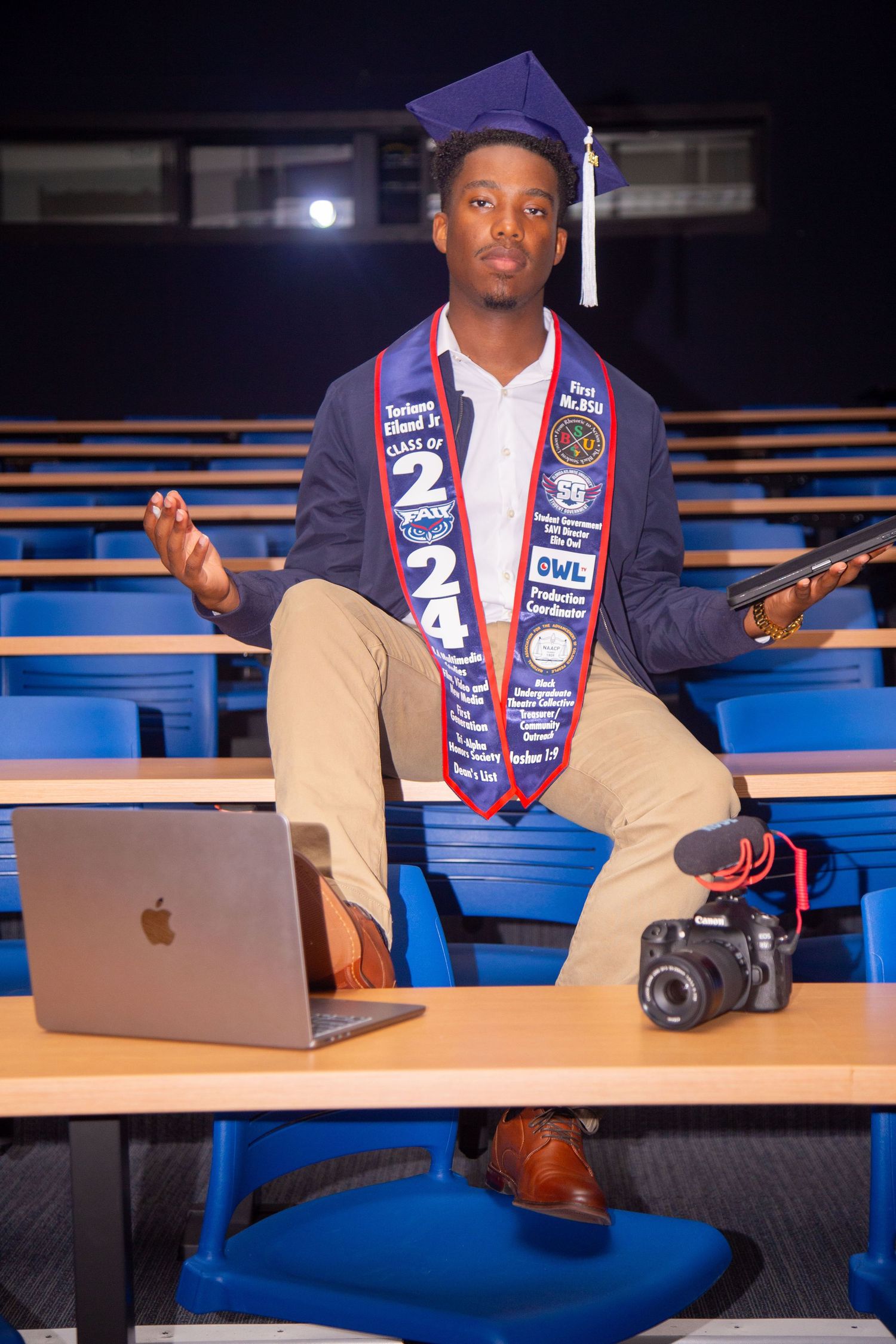 Graduate poses with laptop and camera on bleachers wearing graduation regalia.