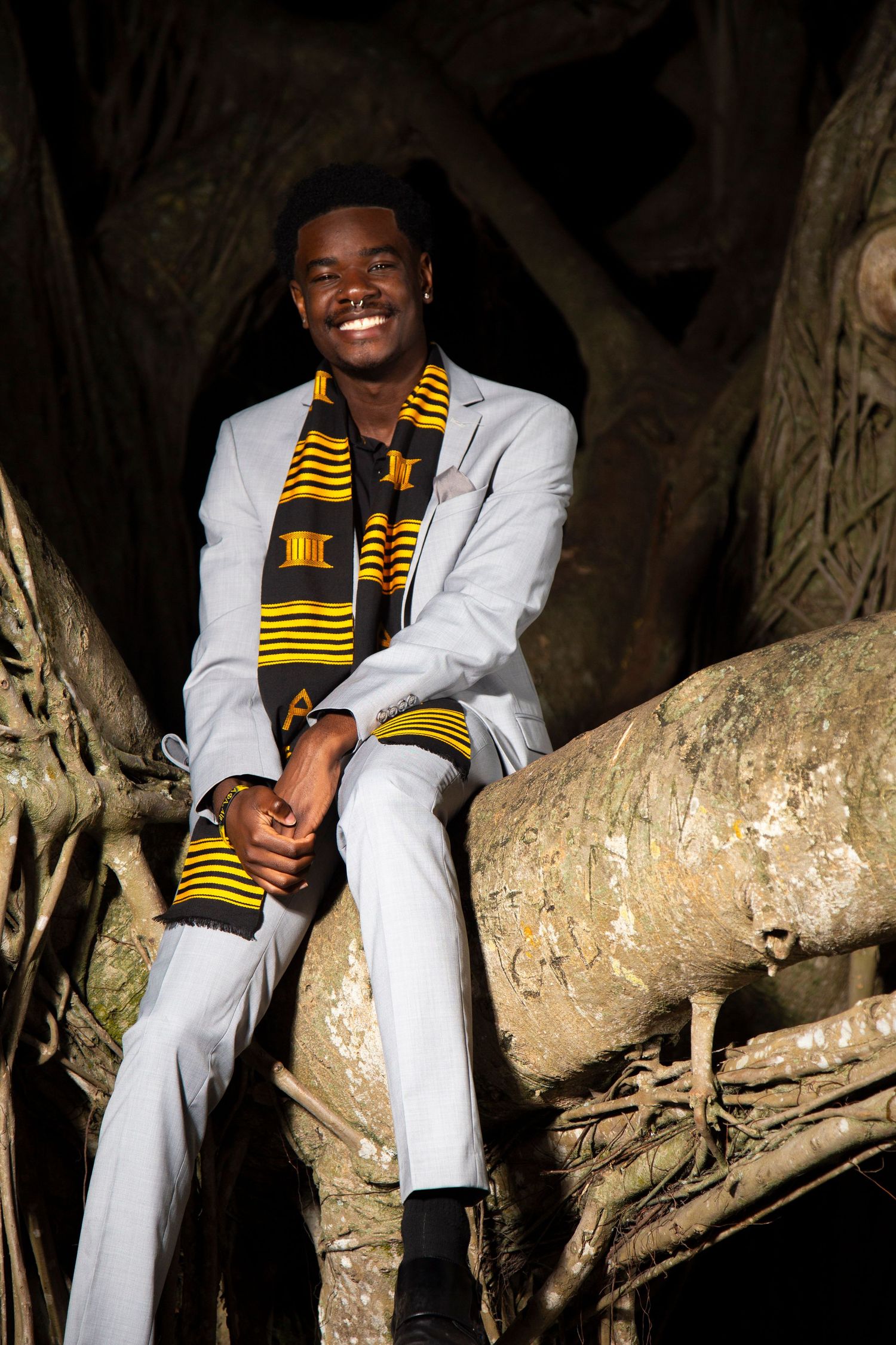 Graduate in white suit with kente stole sits on a large tree branch at night.