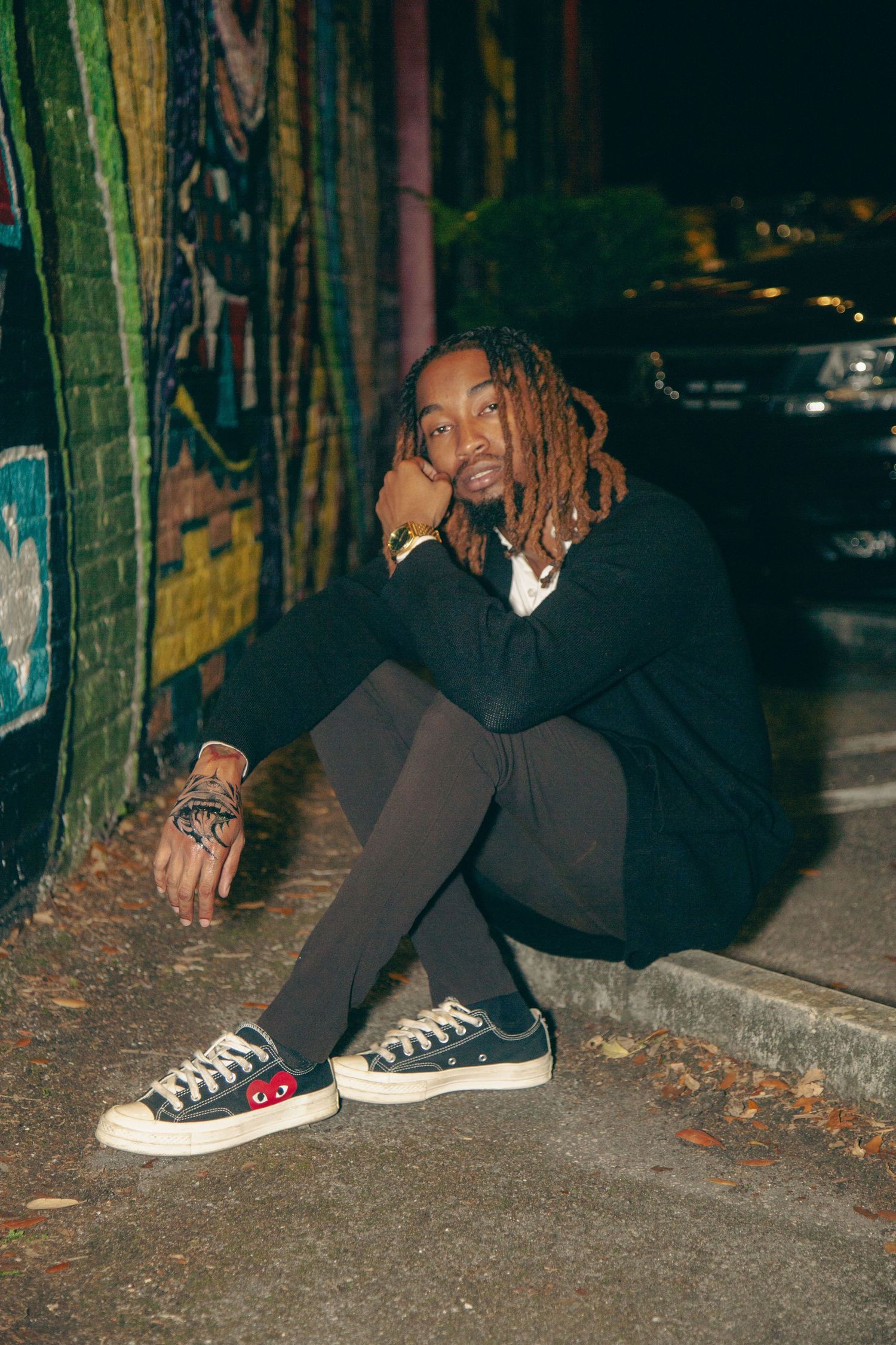 Person in black outfit sits casually on curb wearing Converse sneakers against graffiti wall.
