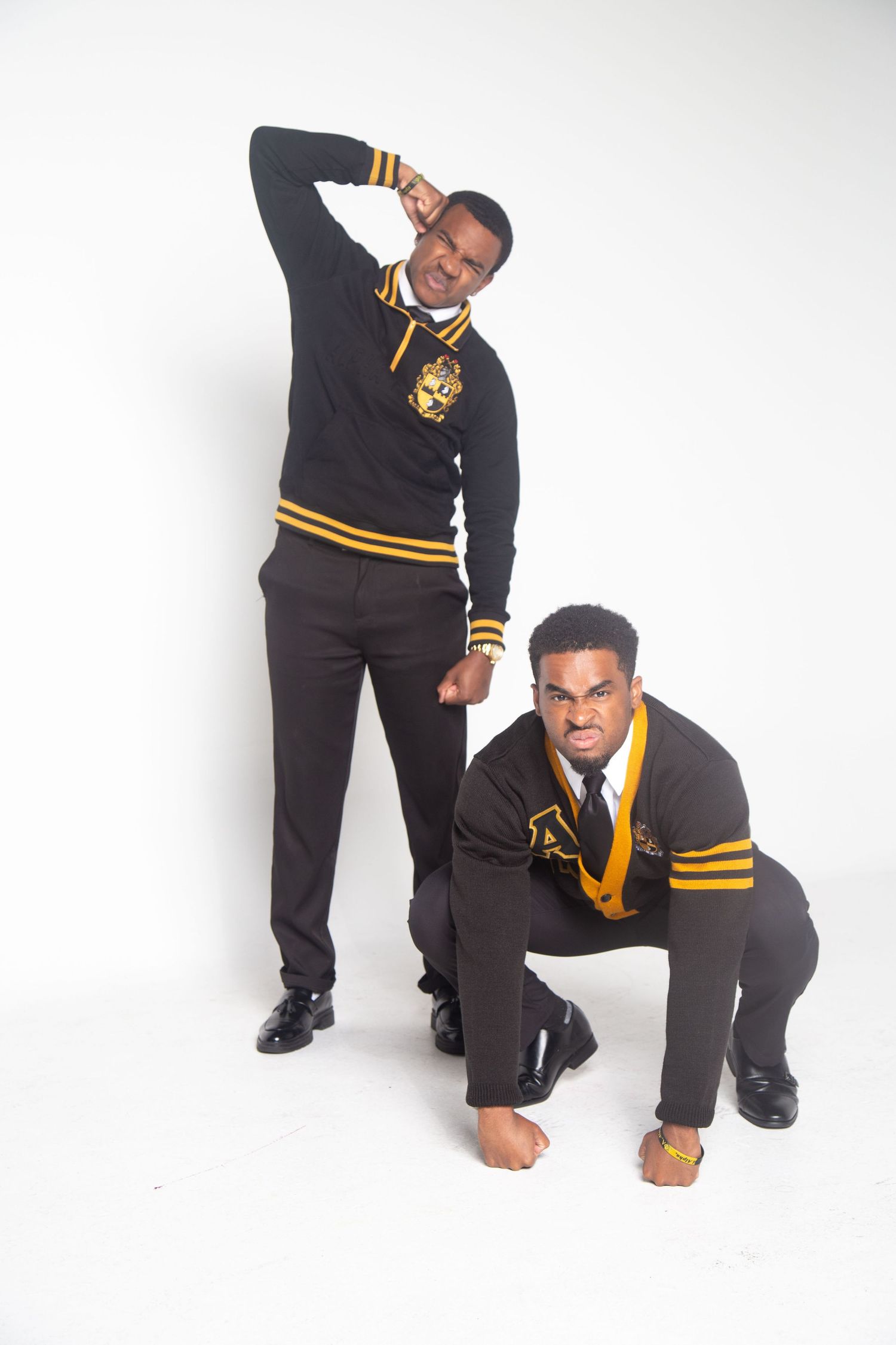 Two people in black and gold fraternity sweaters pose dynamically against a white background.