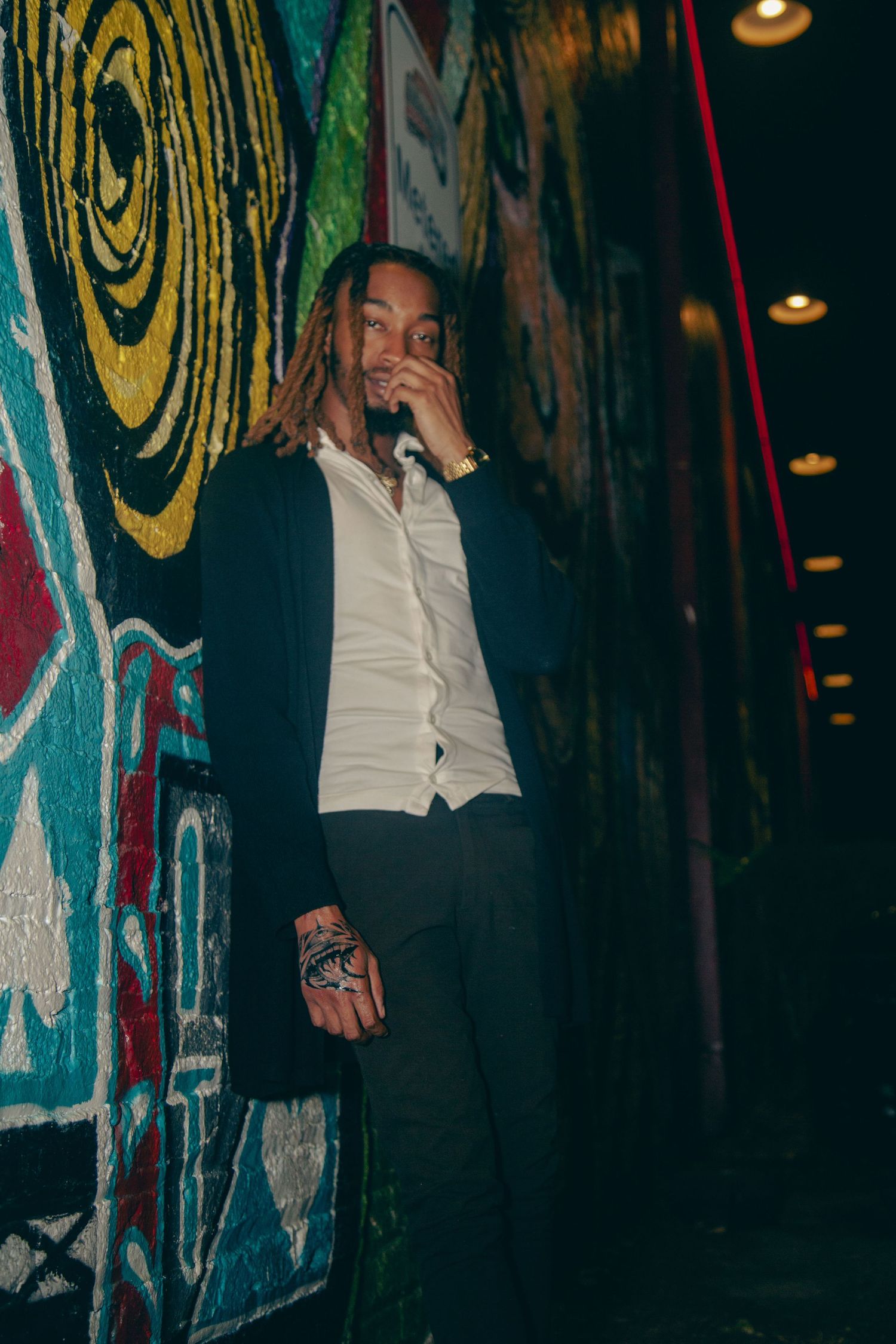 Person in black jacket and white shirt poses against colorful graffiti wall with moody lighting.
