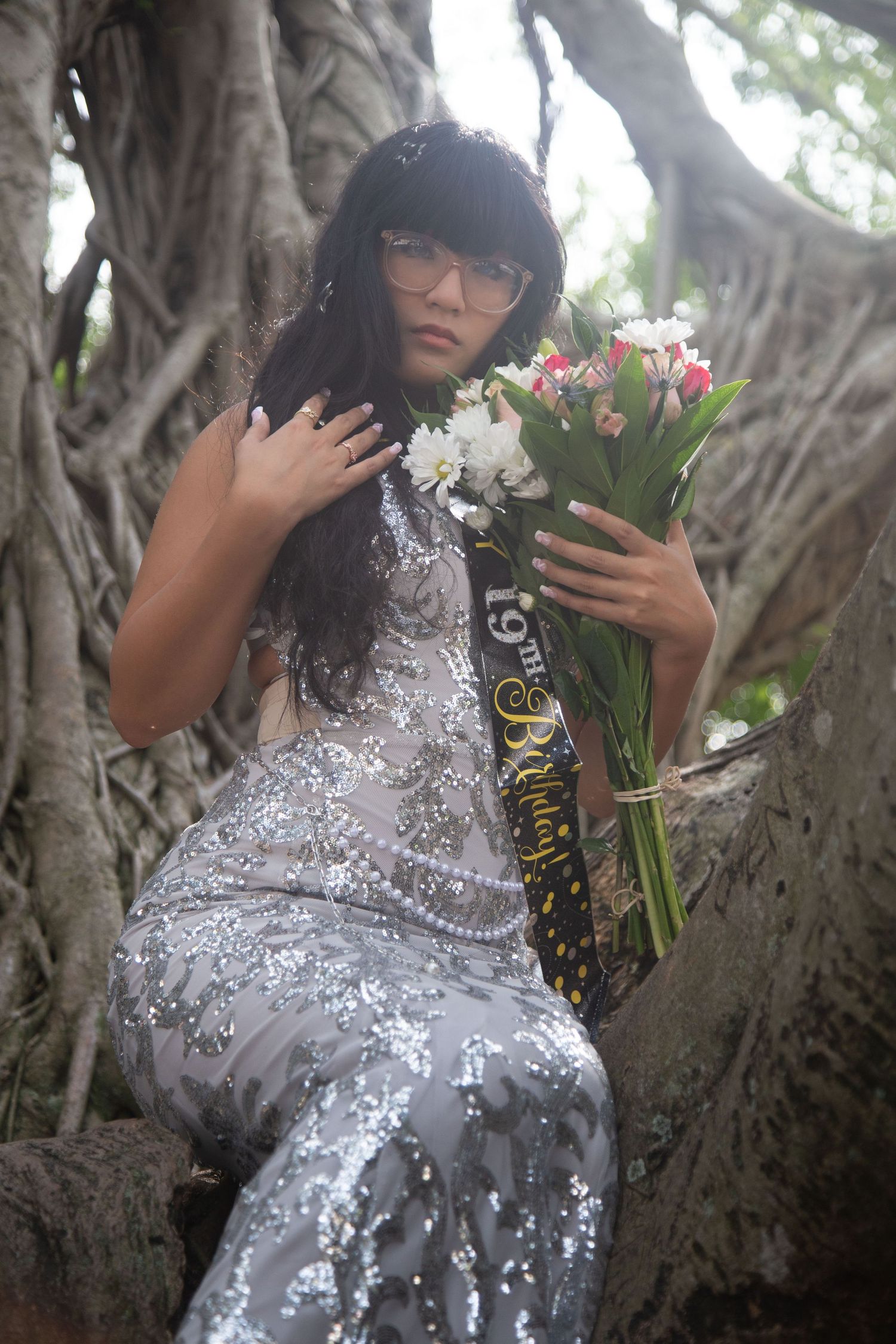Woman in glittering gown holds floral bouquet while posing against banyan tree backdrop.