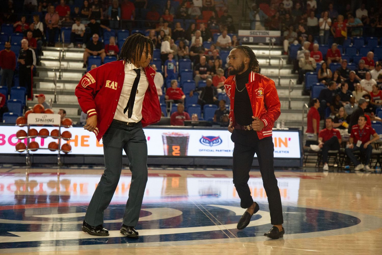 Performers in dark outfits dancing on basketball court during indoor sports event.