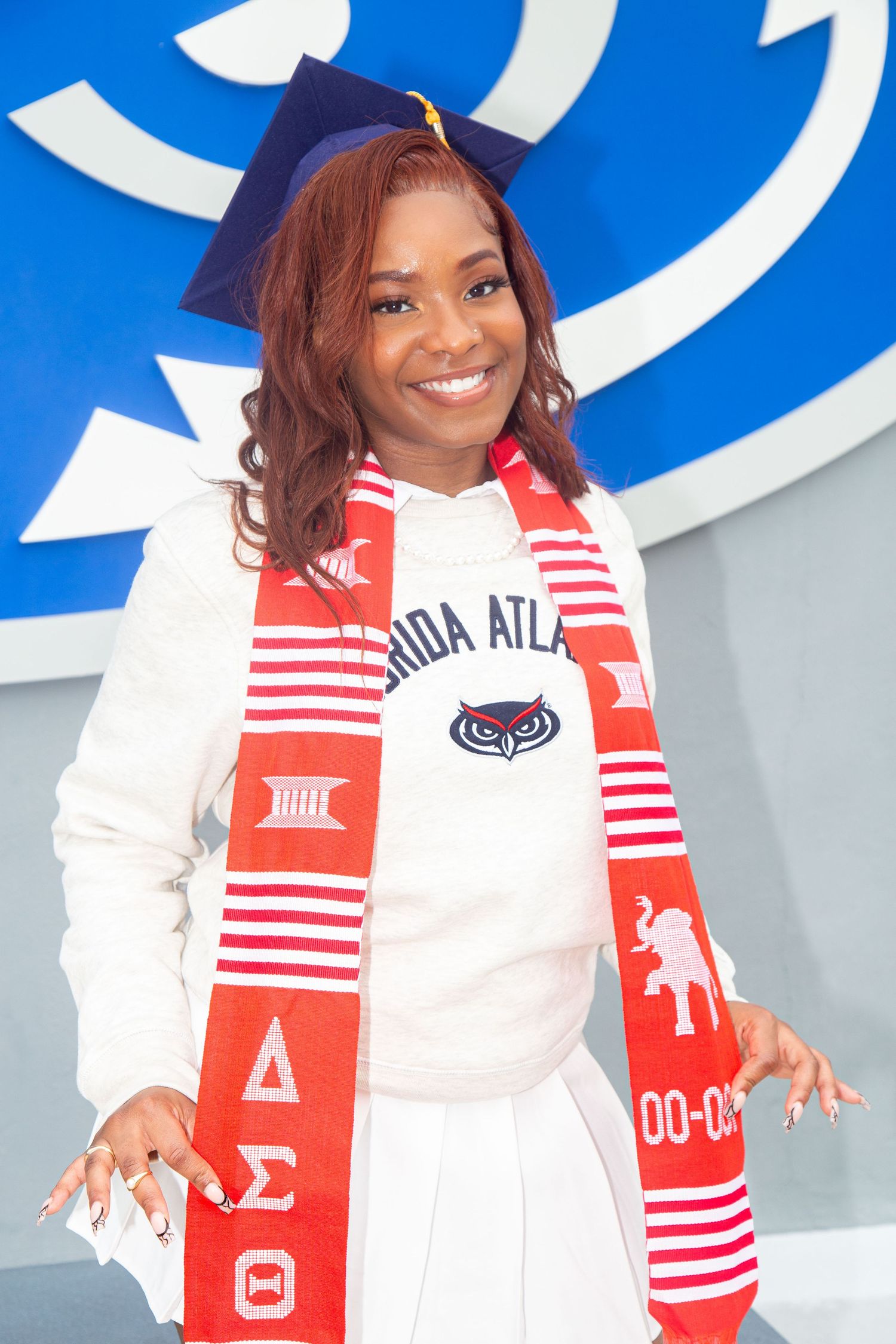 Graduate wearing Delta Sigma Theta sorority scarf poses in front of university logo wall.