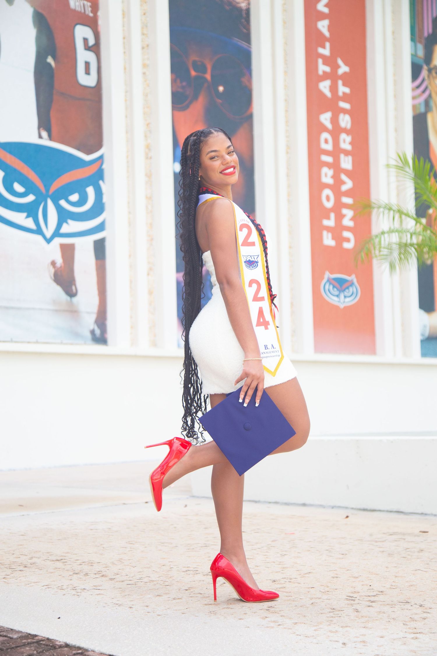 Graduate in white dress and navy graduation gown strikes playful pose on campus steps.