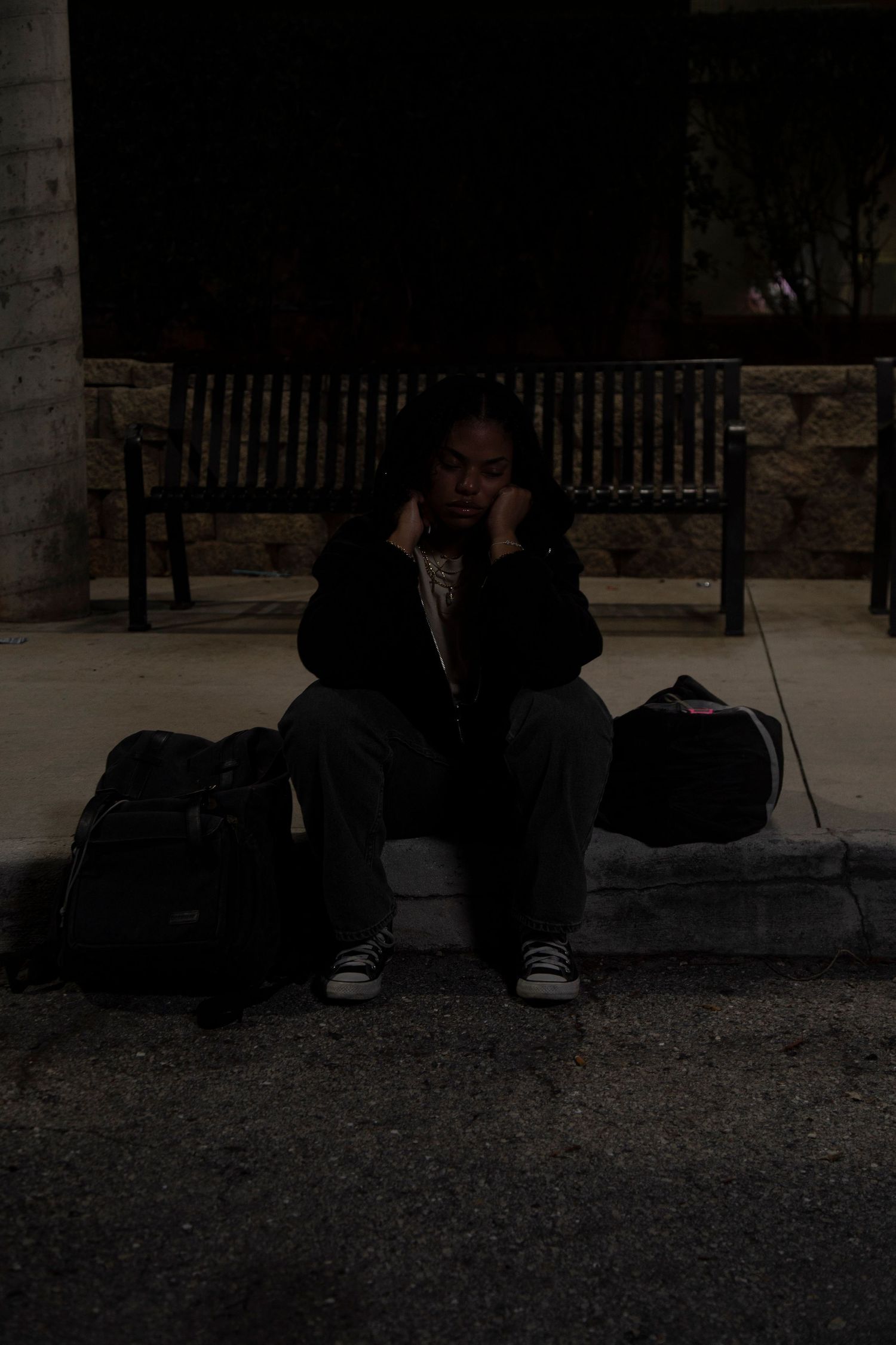 Person sitting alone on curb at night with bags beside them.