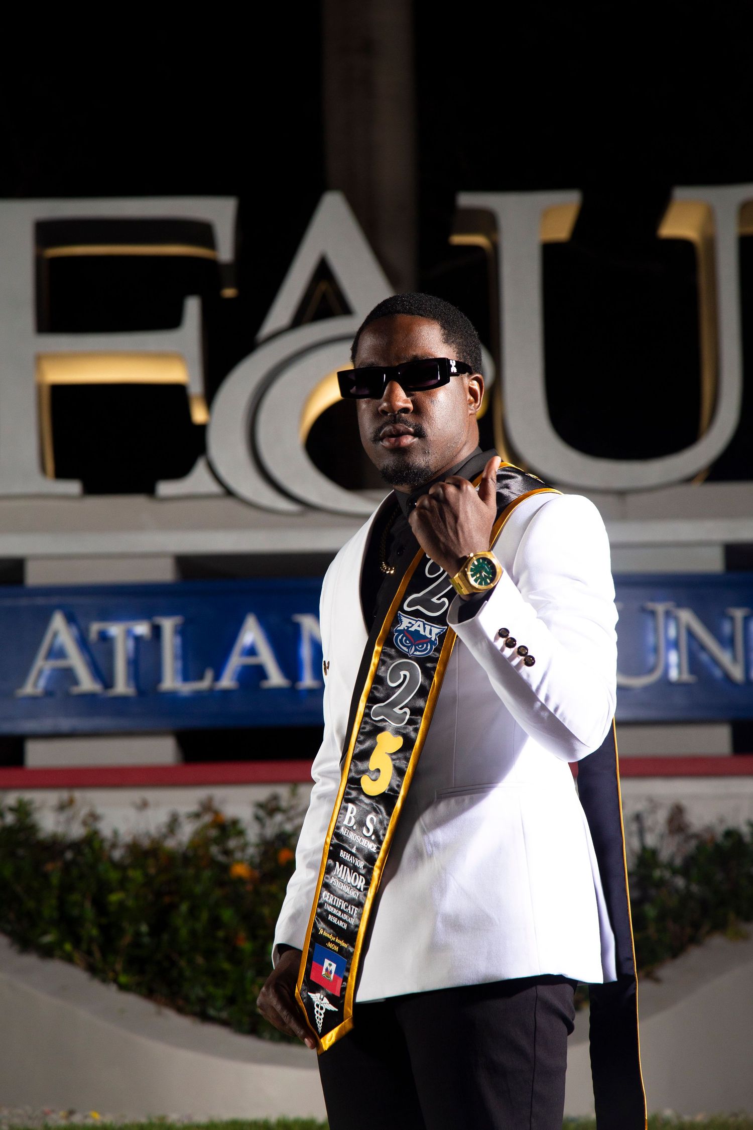 Person in sunglasses and white shirt with decorative sash posing in front of illuminated FIU entrance sign.