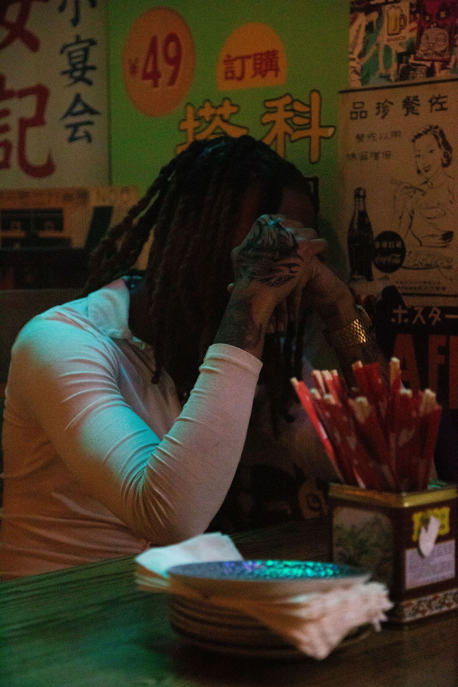 Person with long braids rests head in hands at a restaurant with Chinese characters visible on wall.