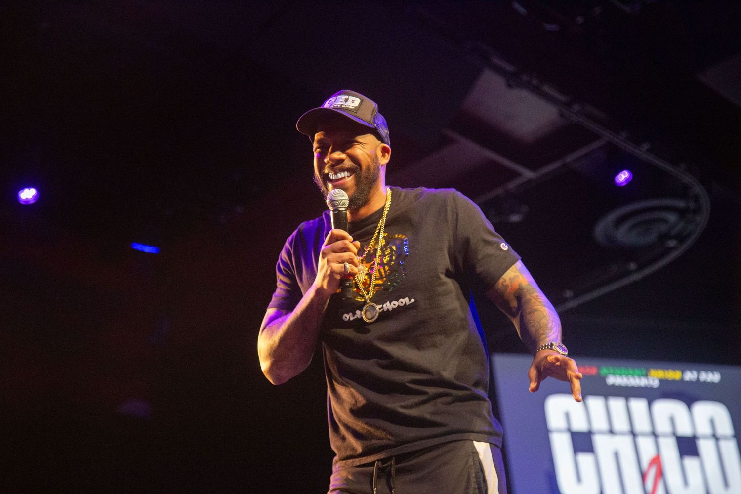 Performer in baseball cap and dark shirt entertains crowd on stage.