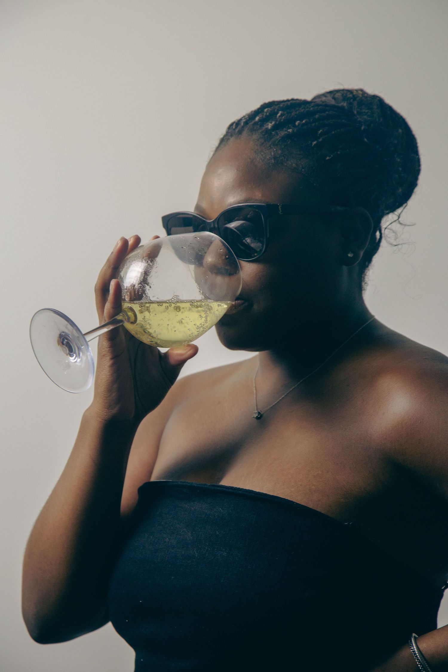 Stylish person in black strapless top and sunglasses sips from a wine glass containing yellow liquid.