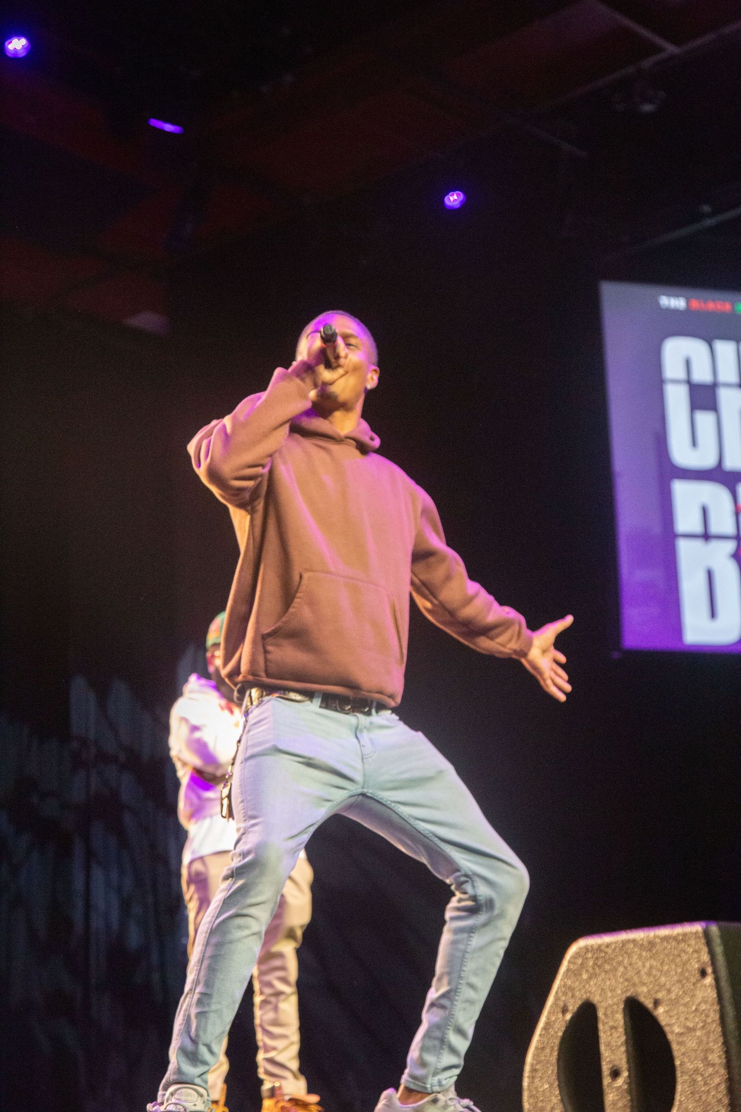 Performer in hoodie and jeans sings on stage with purple lighting.