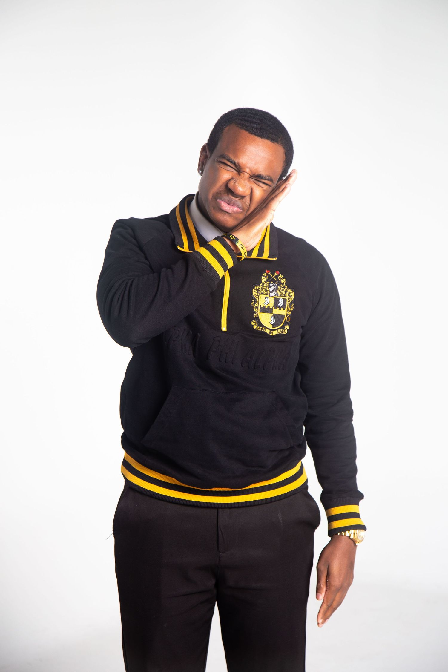 Person in black and gold fraternity sweater poses with hand on neck against white background.