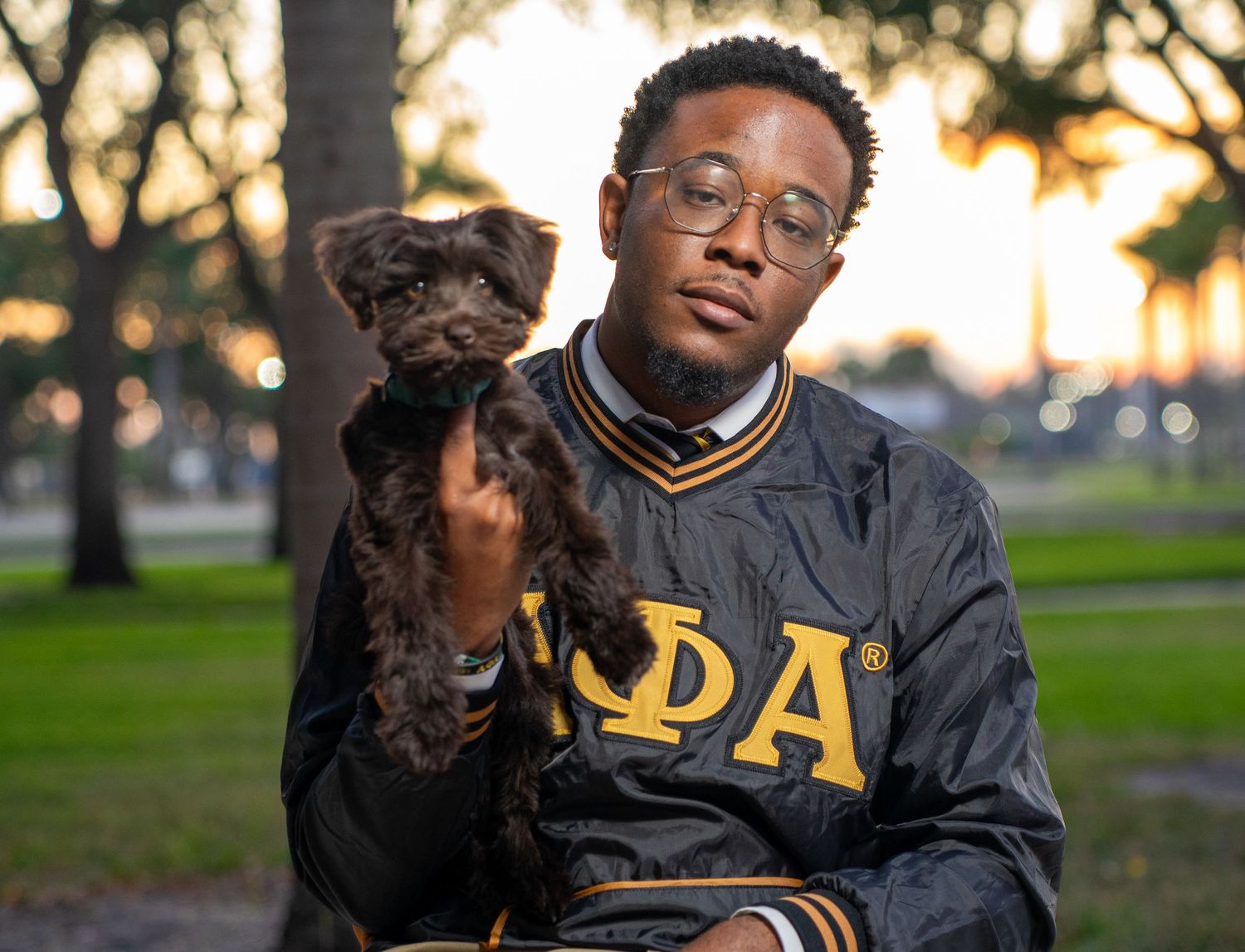 Person in varsity jacket holding black dog outdoors at sunset.