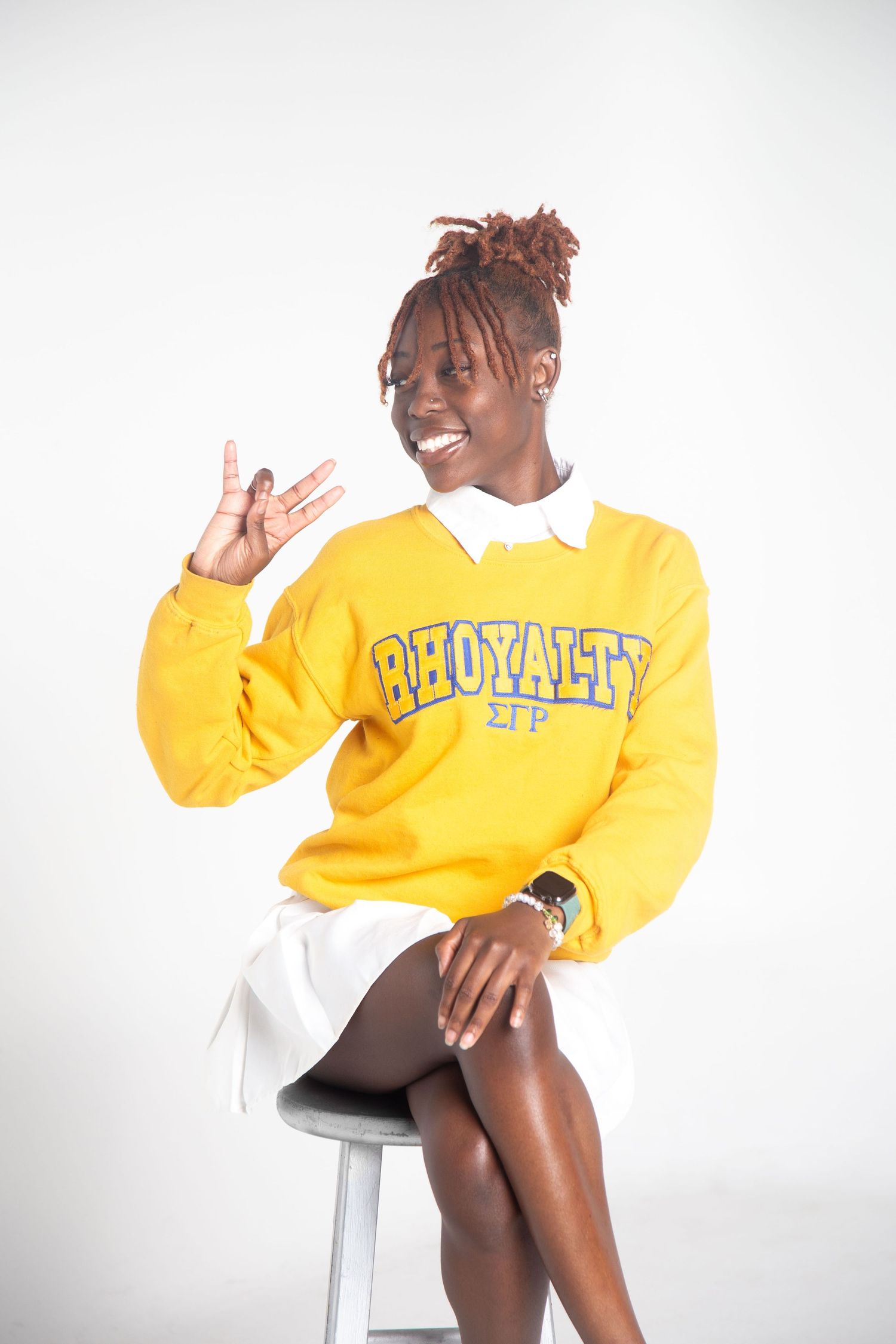 Student wearing bright yellow sorority sweater and white skirt poses cheerfully on stool against white backdrop.