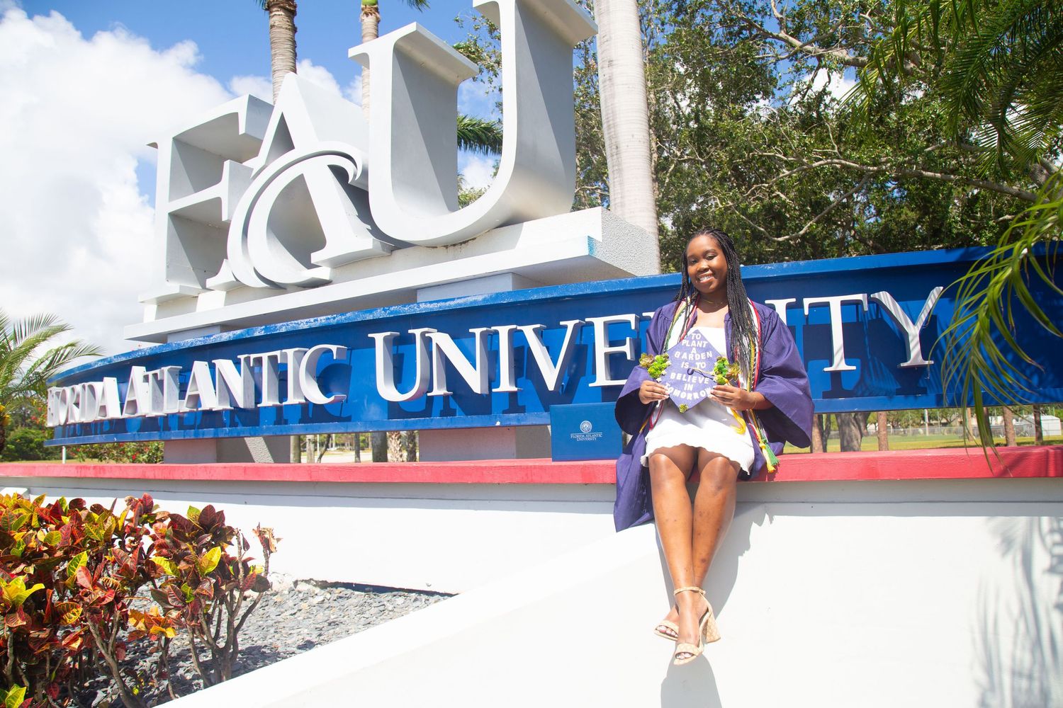 Graduate sits atop the FAU entrance sign in white dress and graduation regalia.