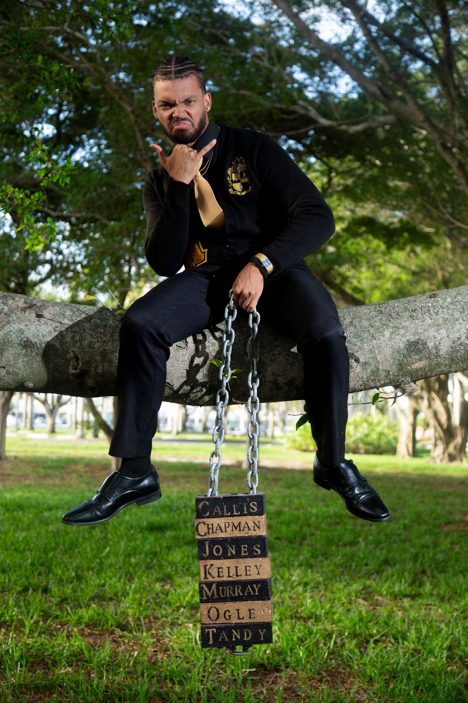 Graduate in black formal attire poses dramatically on tree branch with hanging name plates.