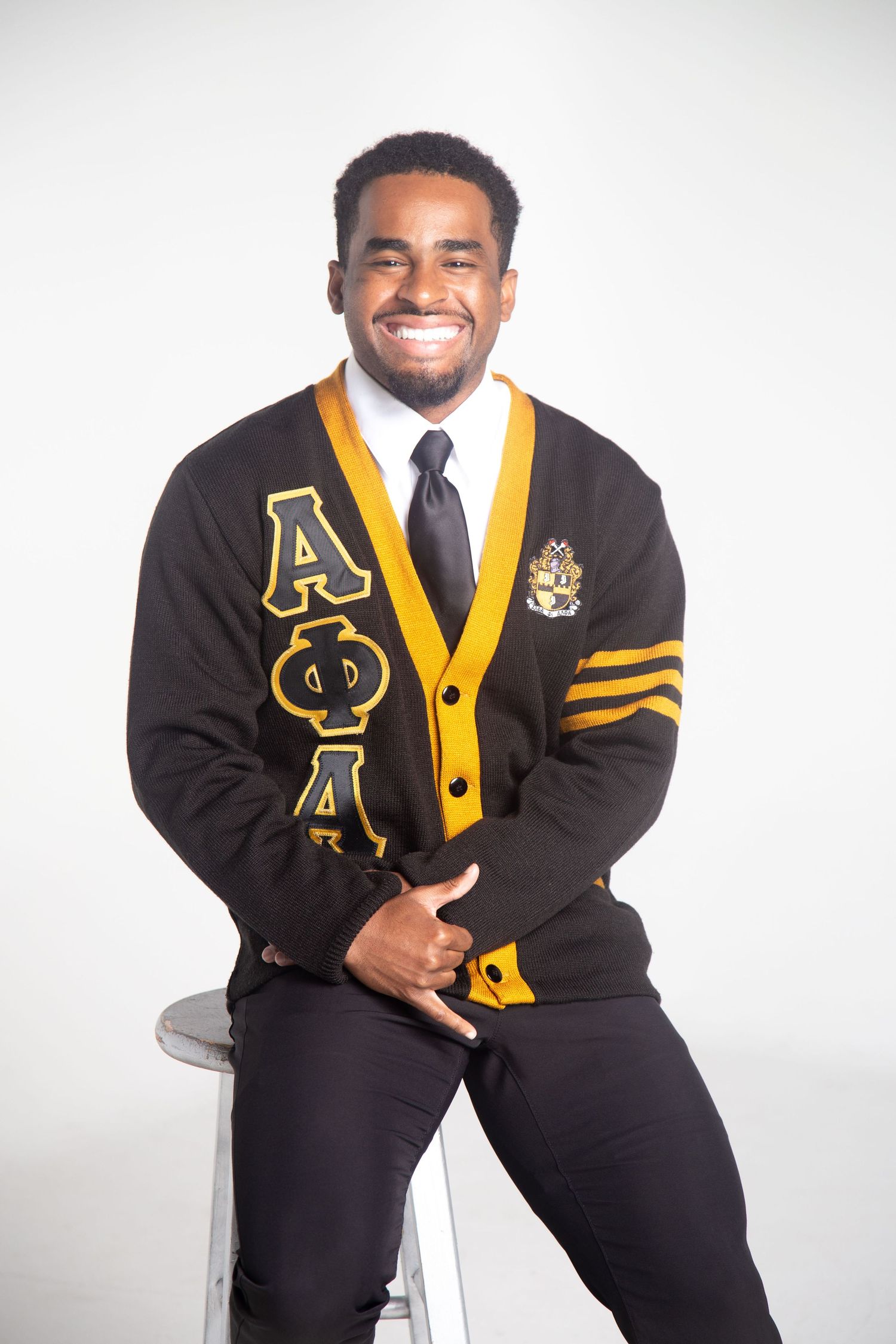 Person in black and gold fraternity cardigan poses on stool in studio setting.