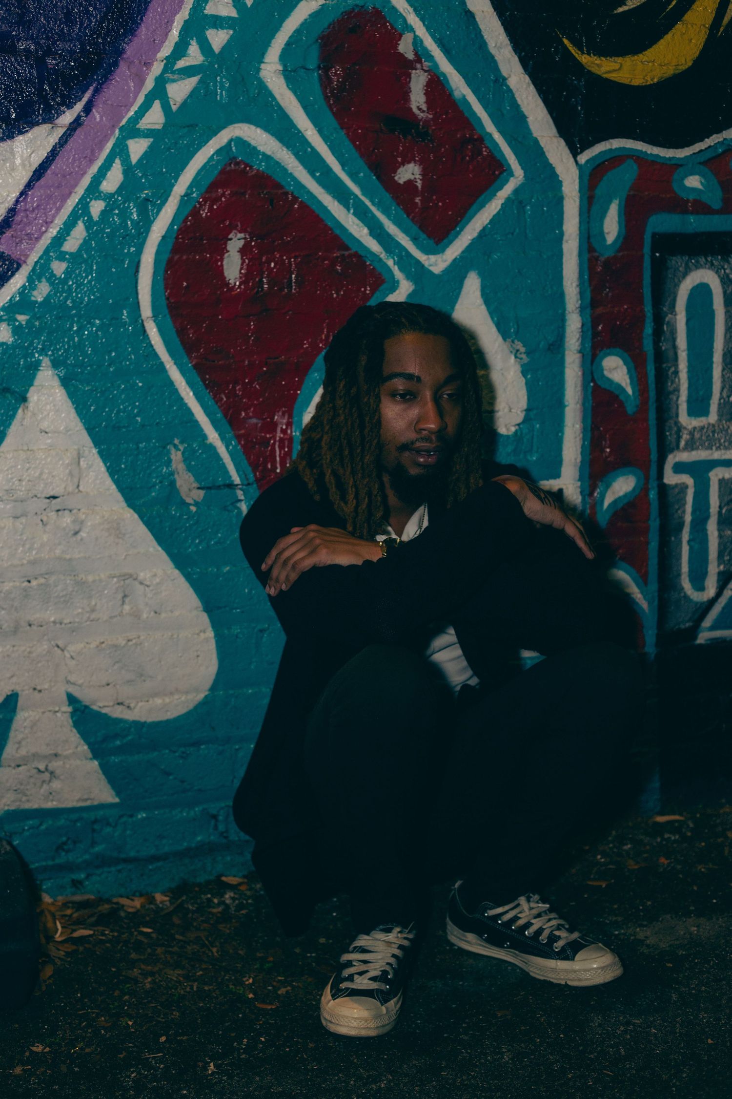 Person crouching against vibrant turquoise and red graffiti mural wearing dark clothing.