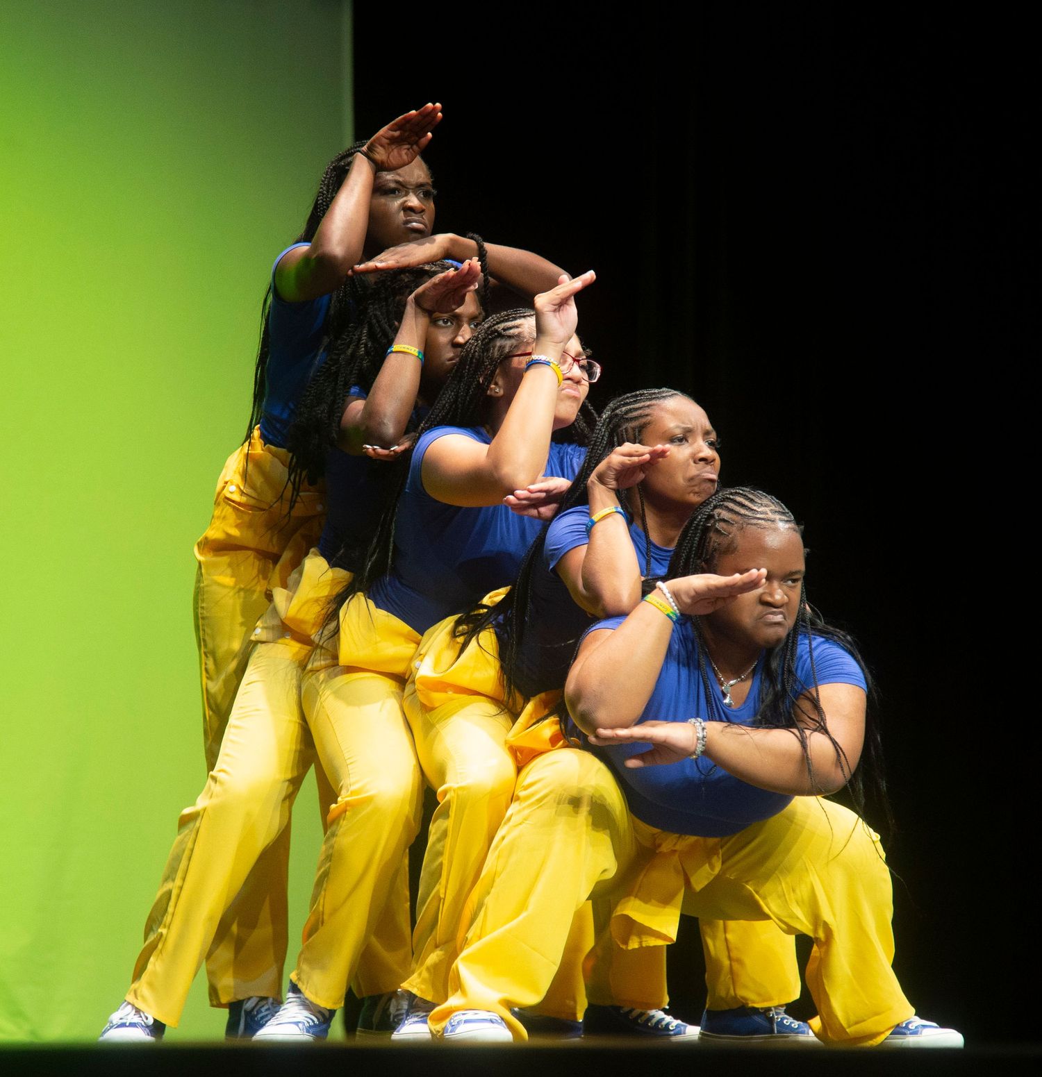 Dance group performs synchronized moves in yellow and blue costumes against green backdrop.
