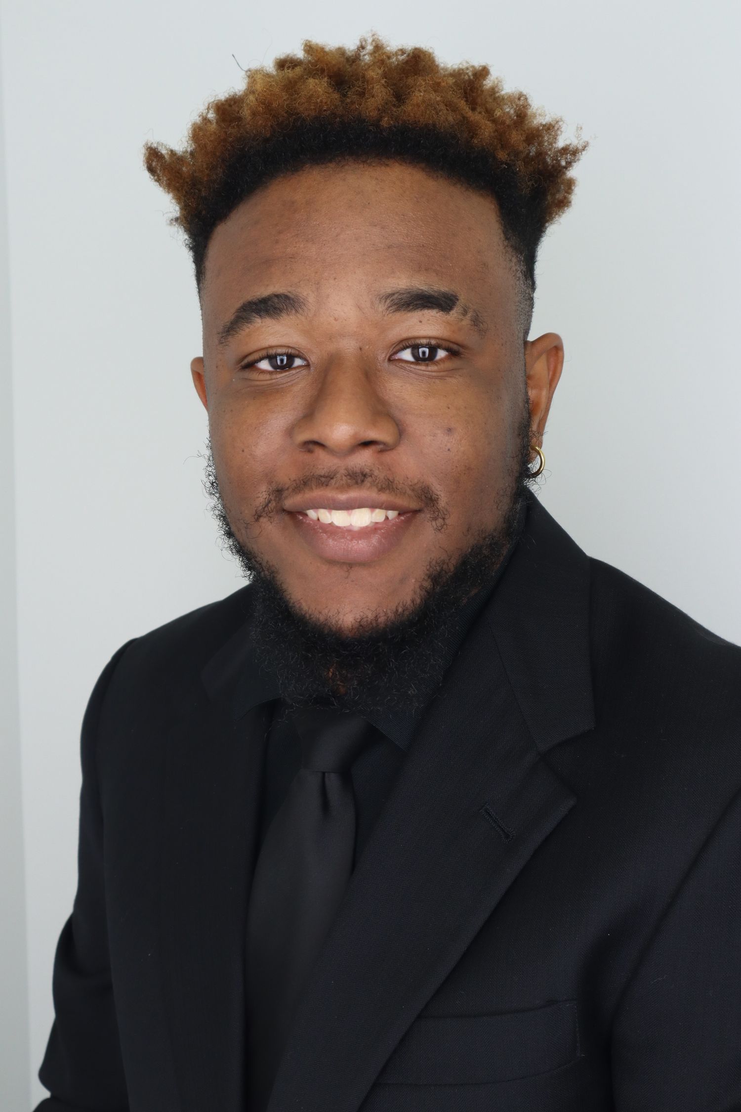 Professional headshot of person in black suit and tie against gray background.