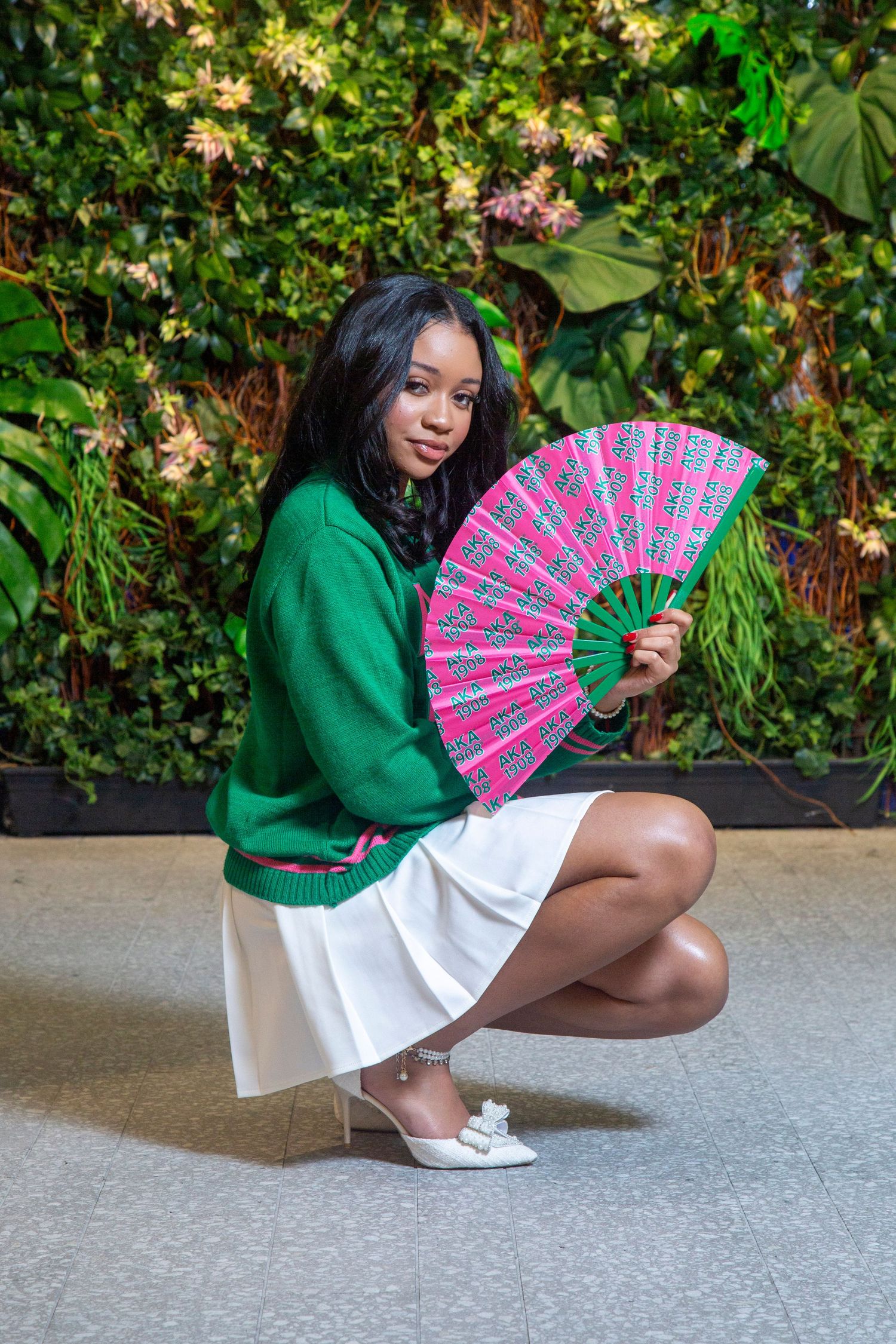 A person in a green sweater and white skirt poses with a pink and white decorative fan against a leafy backdrop.
