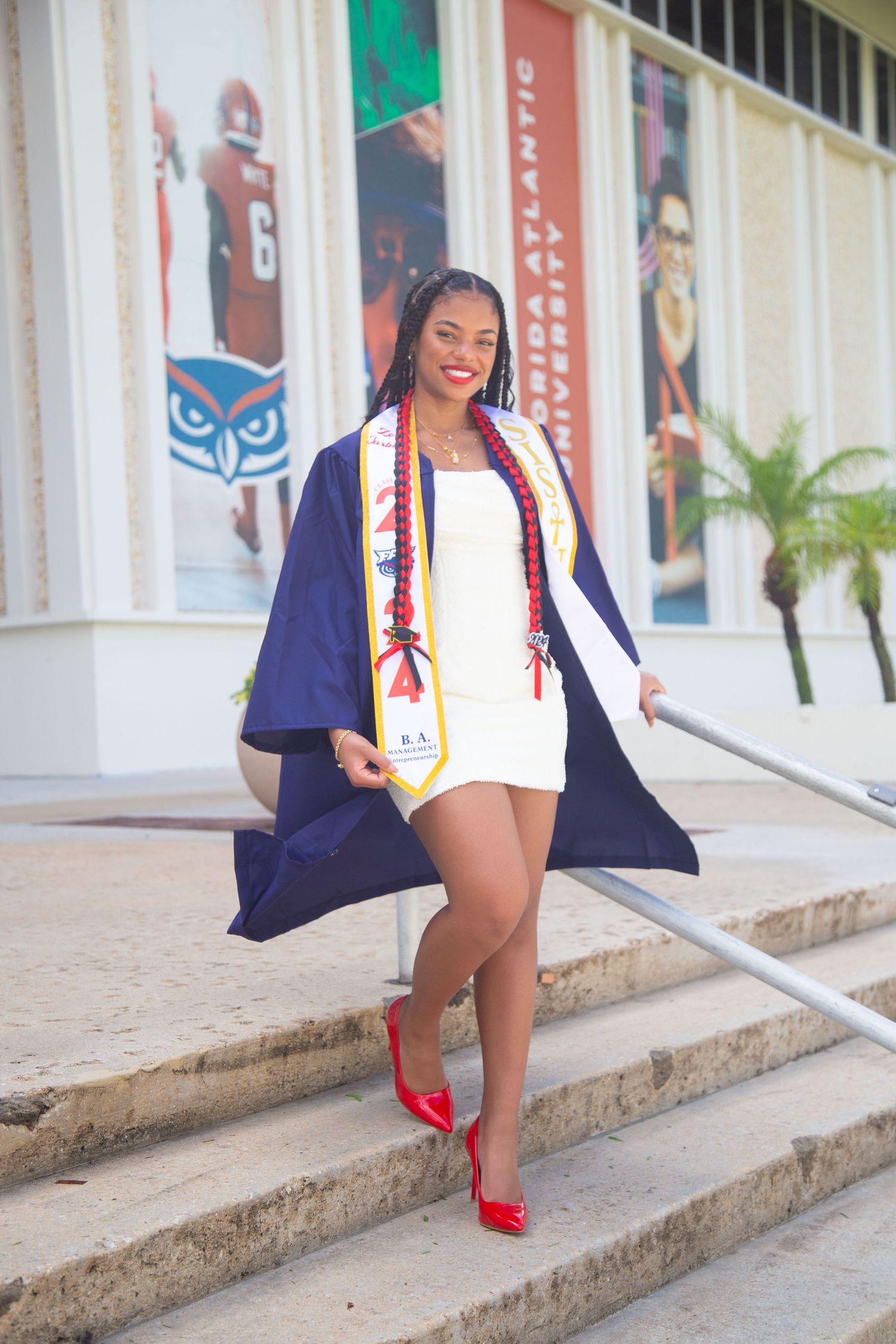 Graduate in academic regalia poses on steps wearing red heels and graduation stole.
