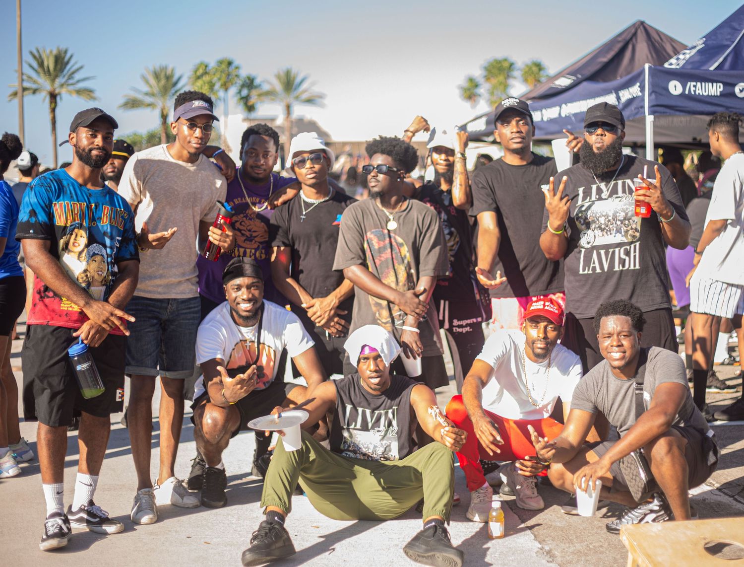 A group of people gathered outdoors in a casual setting with palm trees and tents in the background.