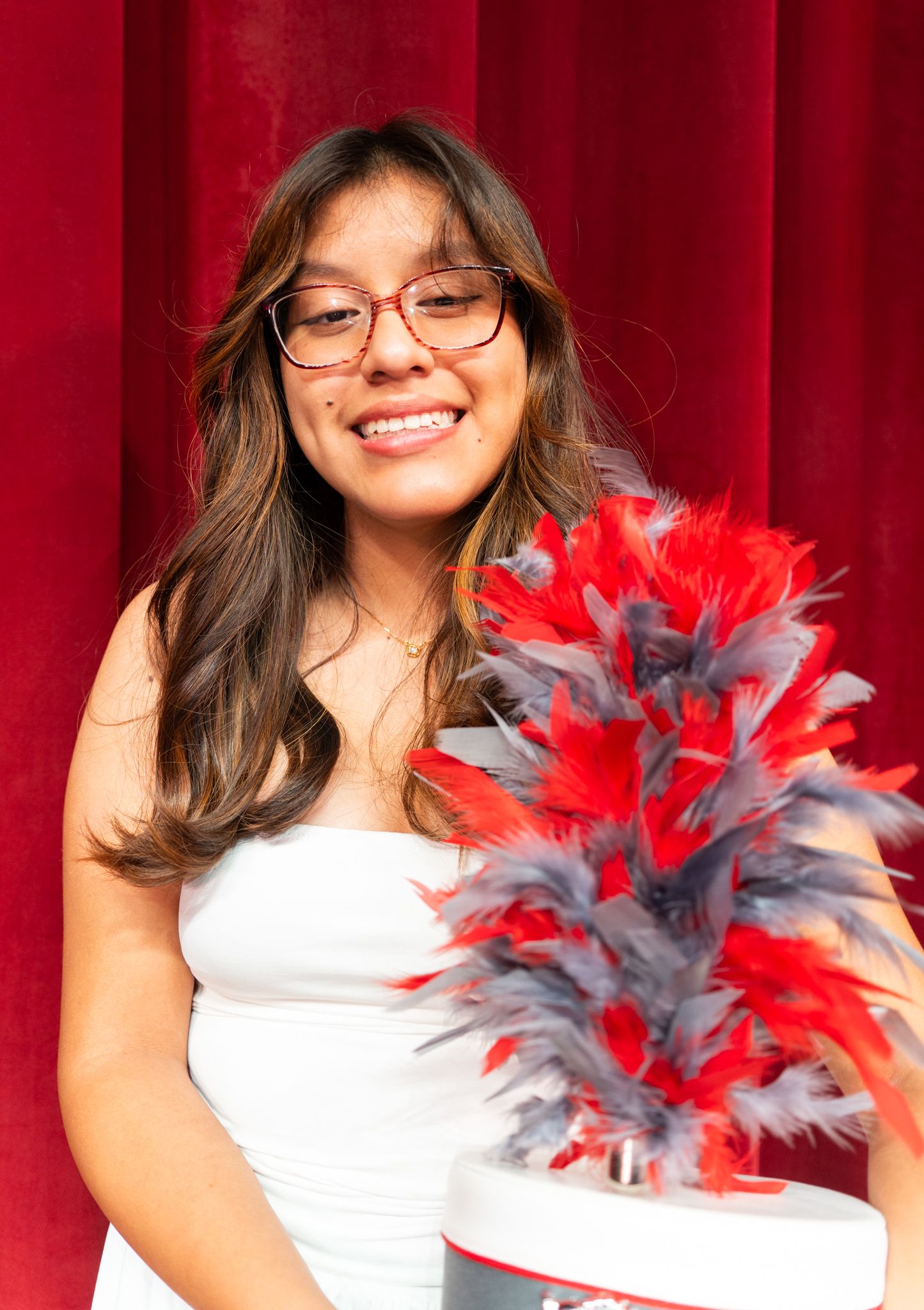 Smiling young woman with glasses in white dress holding a marching band hat with red and gray feathers.