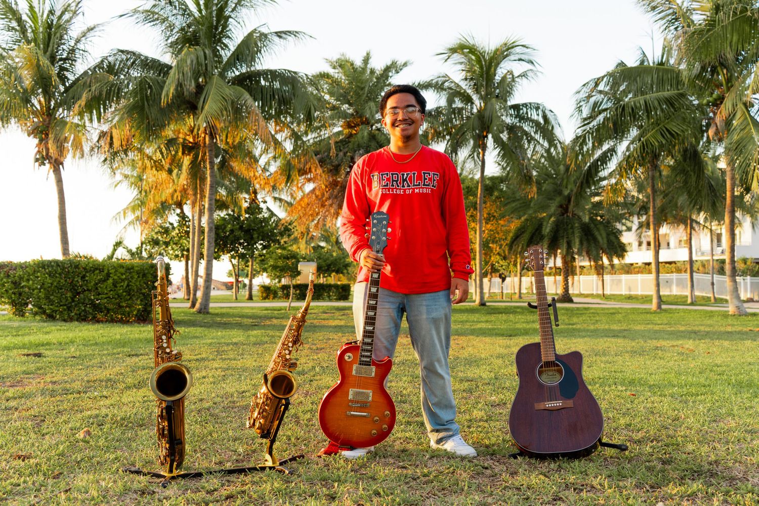 Musician standing outdoors with saxophones and guitars on grass, surrounded by palm trees in a tropical setting.