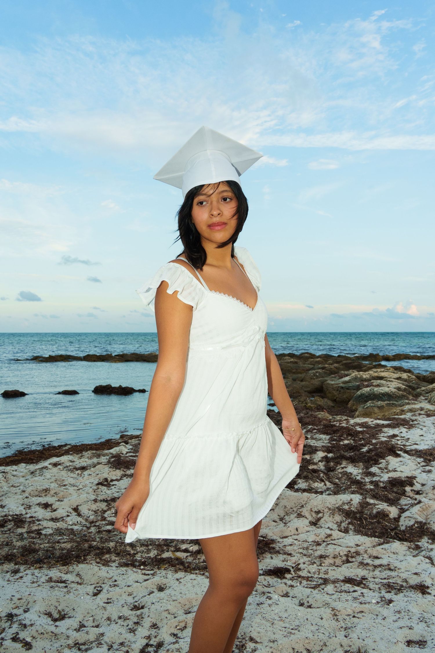 Young woman in white dress and graduation cap poses on rocky beach with ocean and blue sky backdrop.