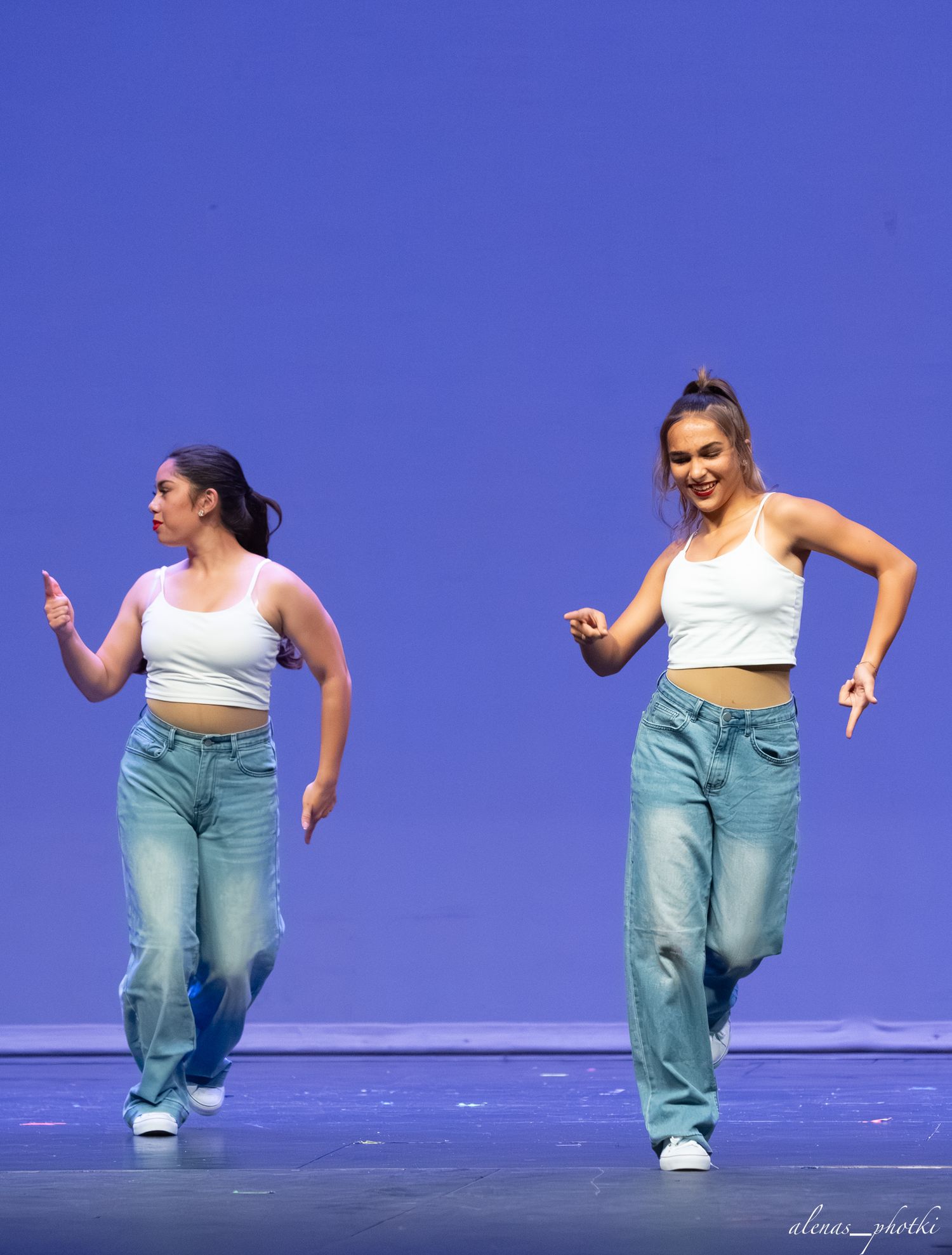 Two women in white crop tops and wide-leg jeans perform a synchronized dance routine on a blue-lit stage.