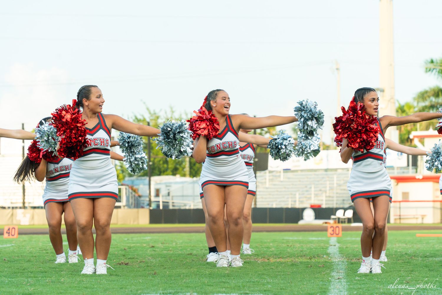 Three cheerleaders in red and white uniforms perform synchronized routine on football field holding metallic pom-poms.