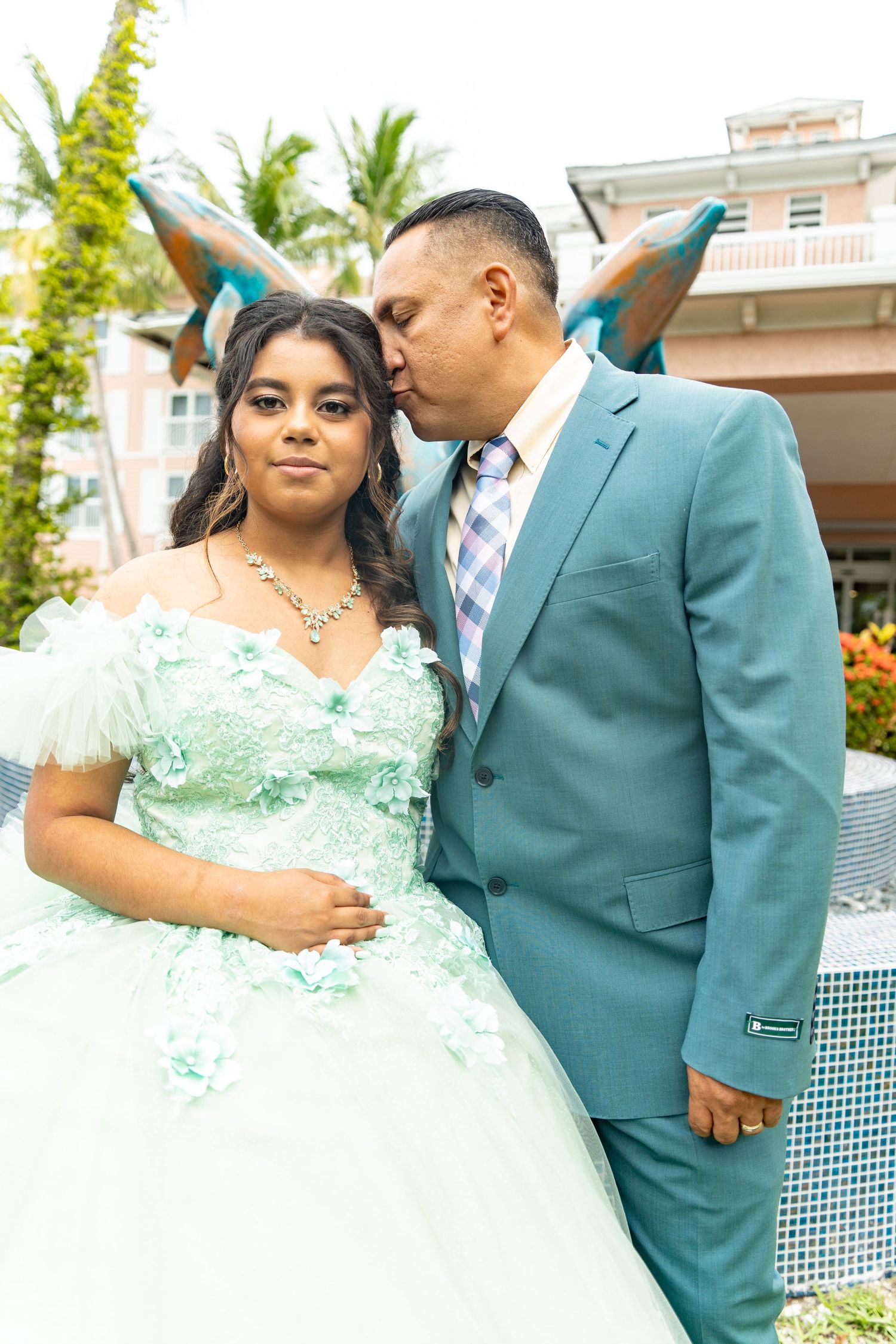 A man in a teal suit kisses a girl in a mint green quinceañera gown outdoors near a fountain.