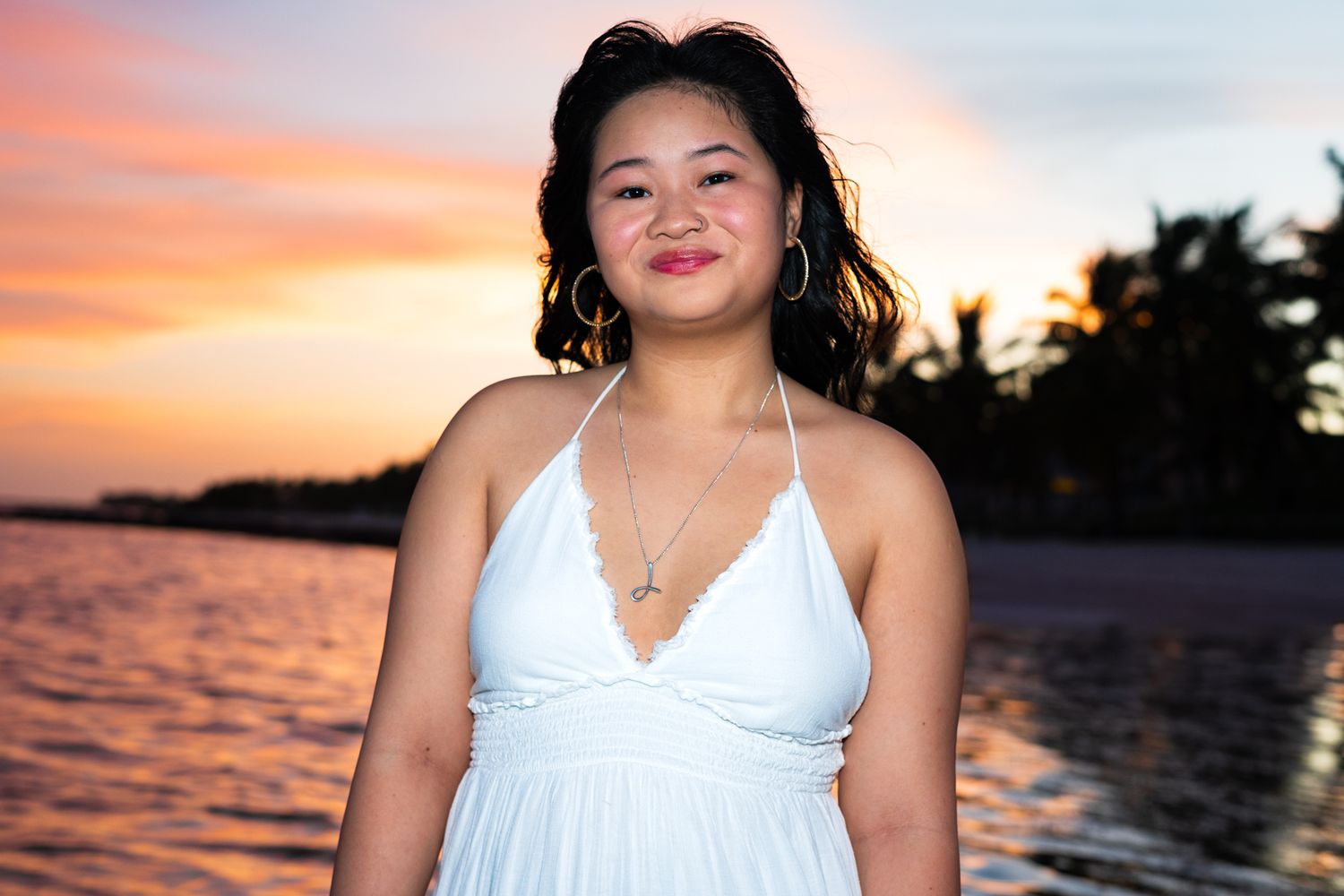 A figure in a white dress poses against a vibrant orange and pink sunset over tropical waters.