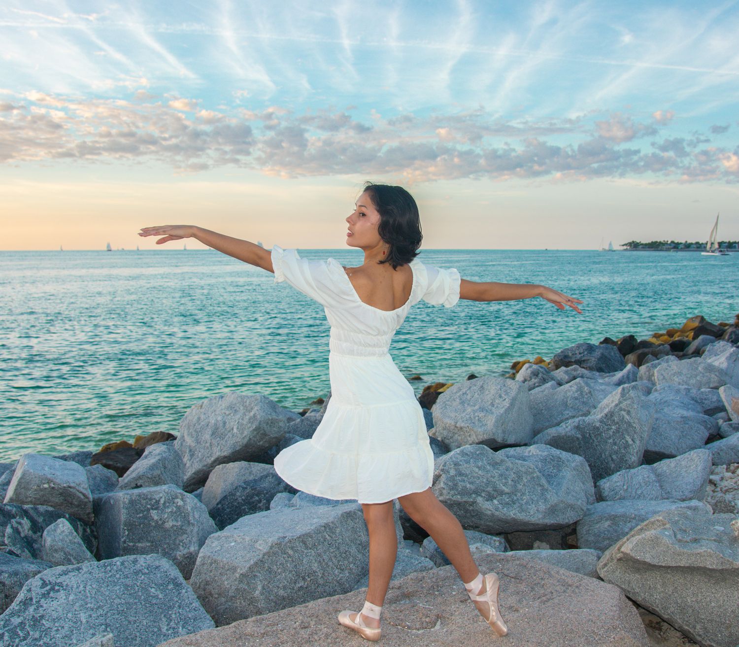 Dancer in white dress performs en pointe on rocky beach at sunset with ocean backdrop.
