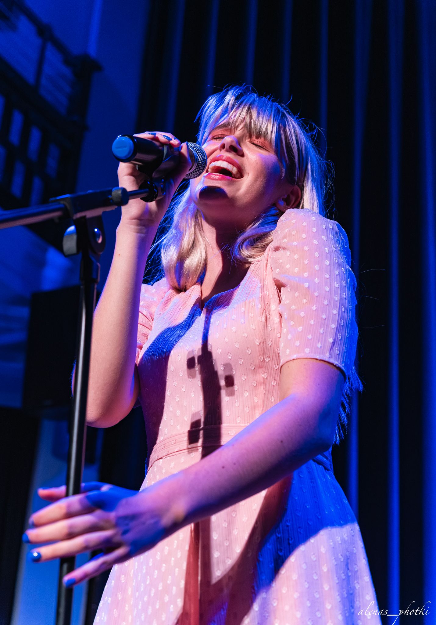 A performer in a polka dot dress sings passionately on stage under blue lighting during a live music performance.